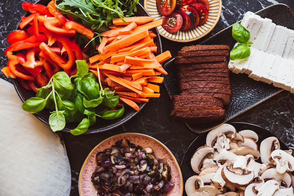 A variety of fresh, sliced vegetables and vegan ingredients on plates, including red bell peppers, carrots, cherry tomatoes, mushrooms, basil, arugula, olives, tofu, and slices of dark bread on a black marble surface.