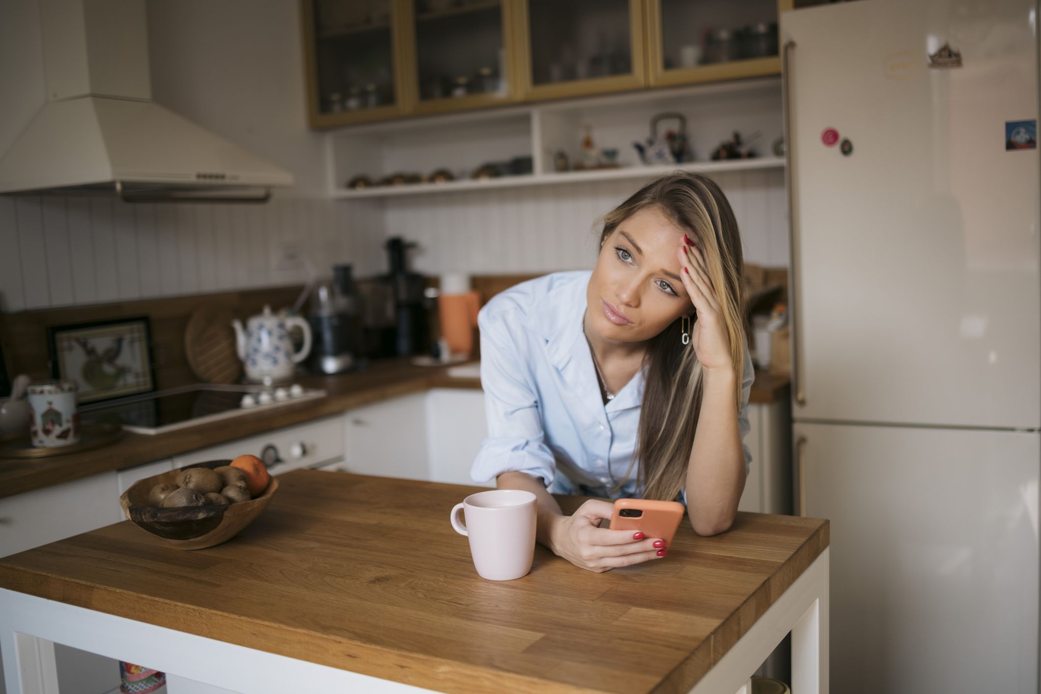 Young woman leaning on kitchen table looking worried while using cell phone. Female with hand on forehead looking away with stressed expressions holding mobile phone in kitchen at home.