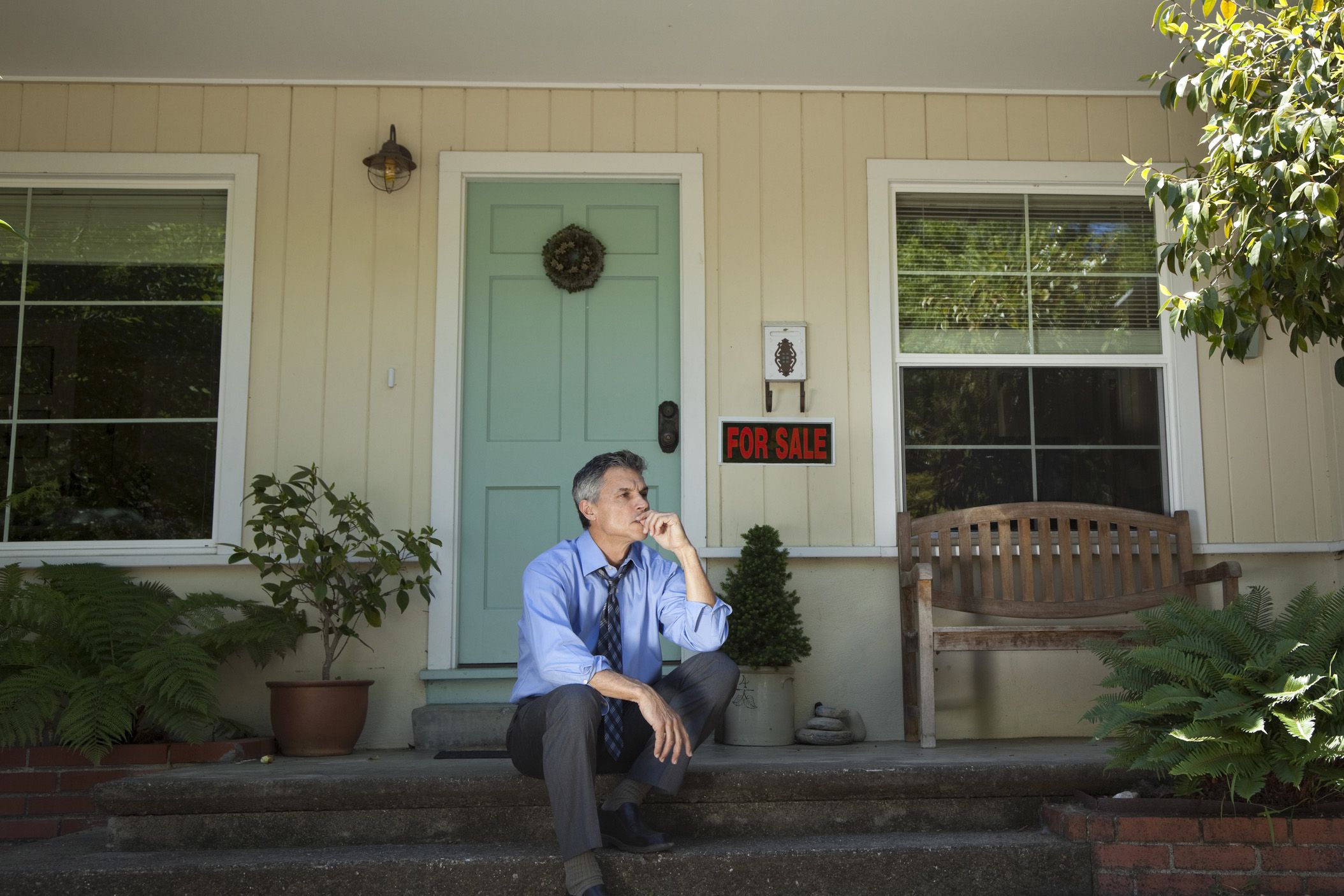 mature man in suit with sad expression sitting outside house with for sale sign