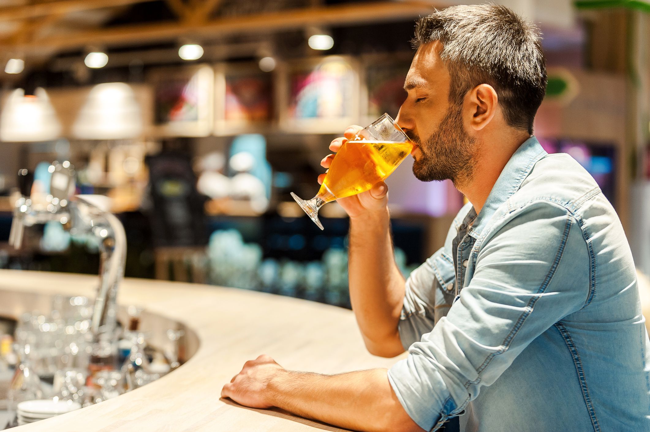 Side view of young man drinking beer and keeping eyes closed while sitting at the bar counter