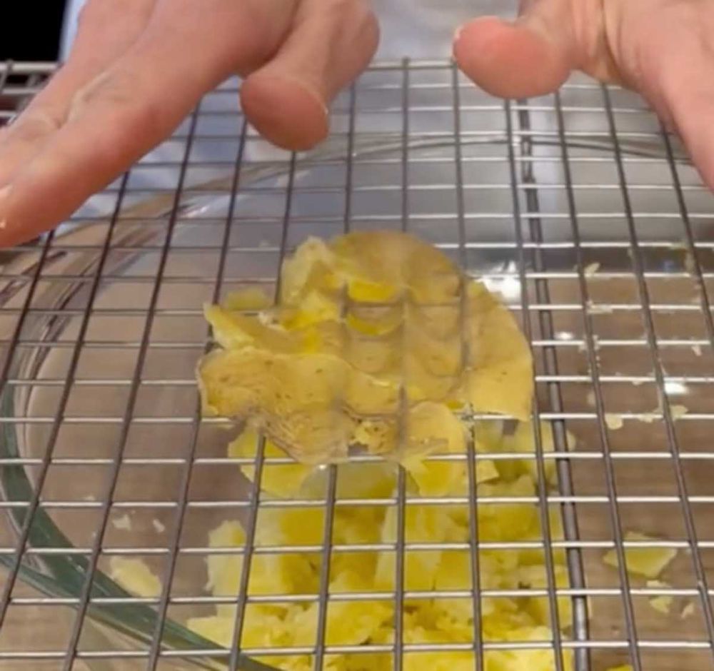 Two hands press a boiled potato through a metal cooling rack over a glass bowl, mashing it into small pieces.