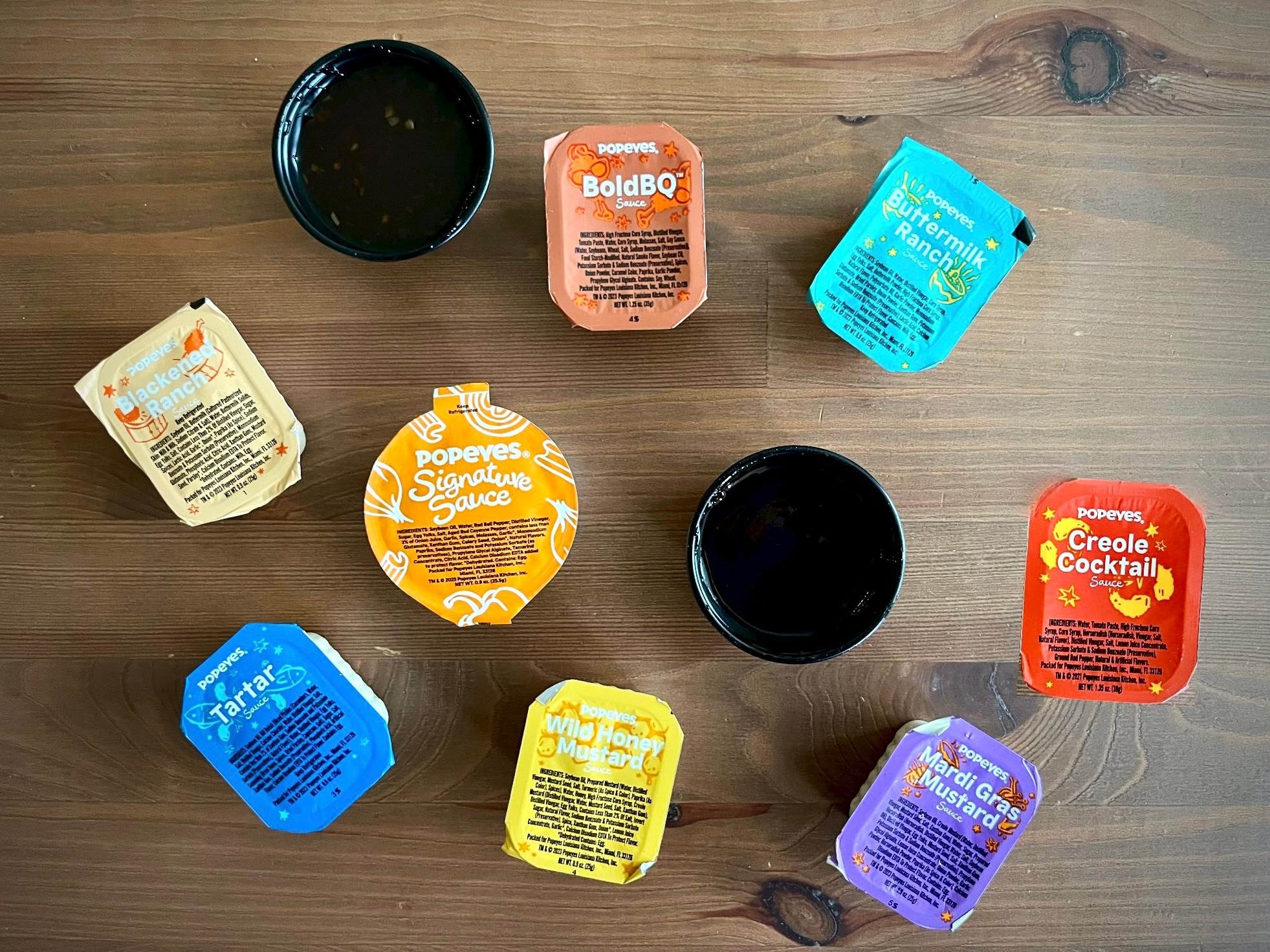 Various Popeyes sauce packets, including BoldBQ, Signature Sauce, Creole Cocktail, Tartar, Wild Honey Mustard, Bayou Buffalo, and Mardi Gras Mustard, are arranged on a wooden table with two open sauce containers.
