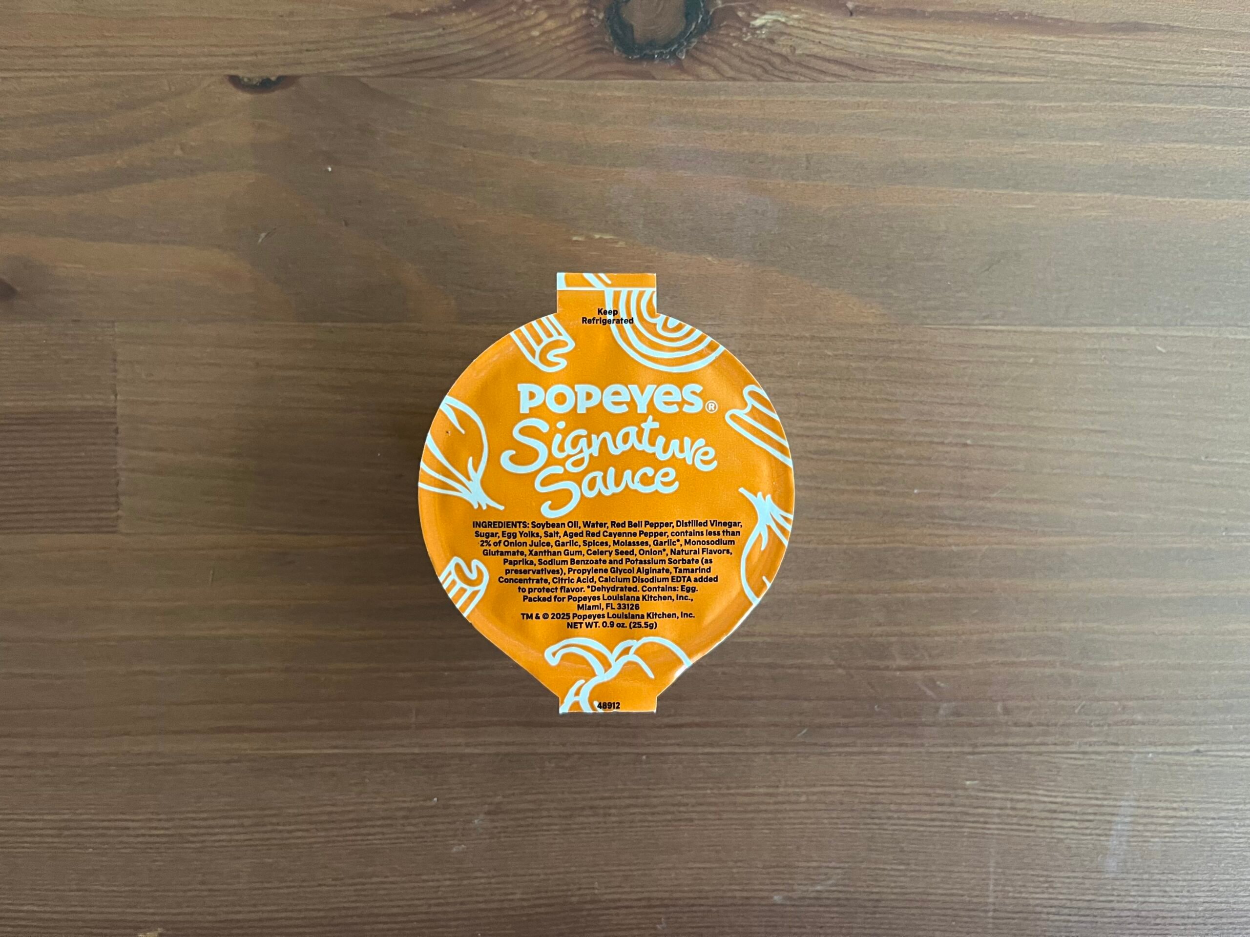A sealed container of Popeyes Signature Sauce sits on a wooden surface. The container is orange with white text and design, displaying the Popeyes logo and sauce name.
