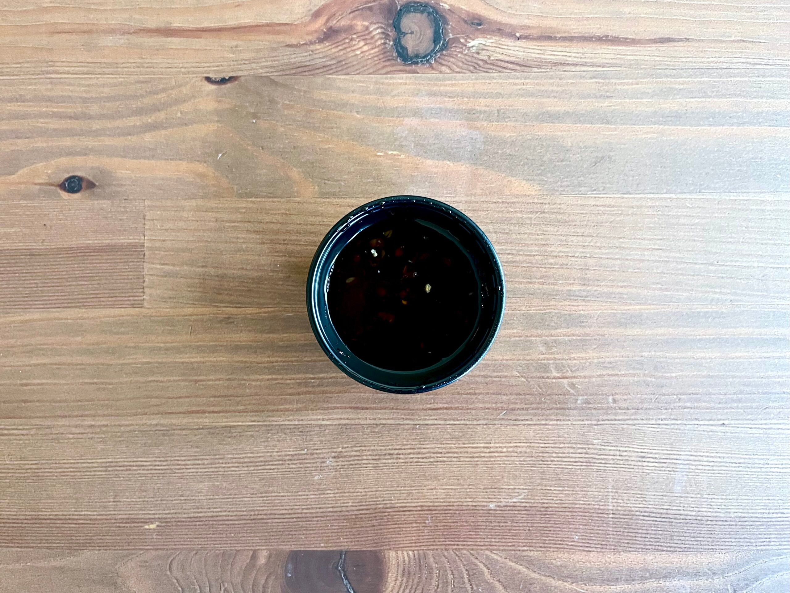 A small black bowl filled with dark sauce, possibly soy sauce, sits on a wooden table with visible grain and knots.