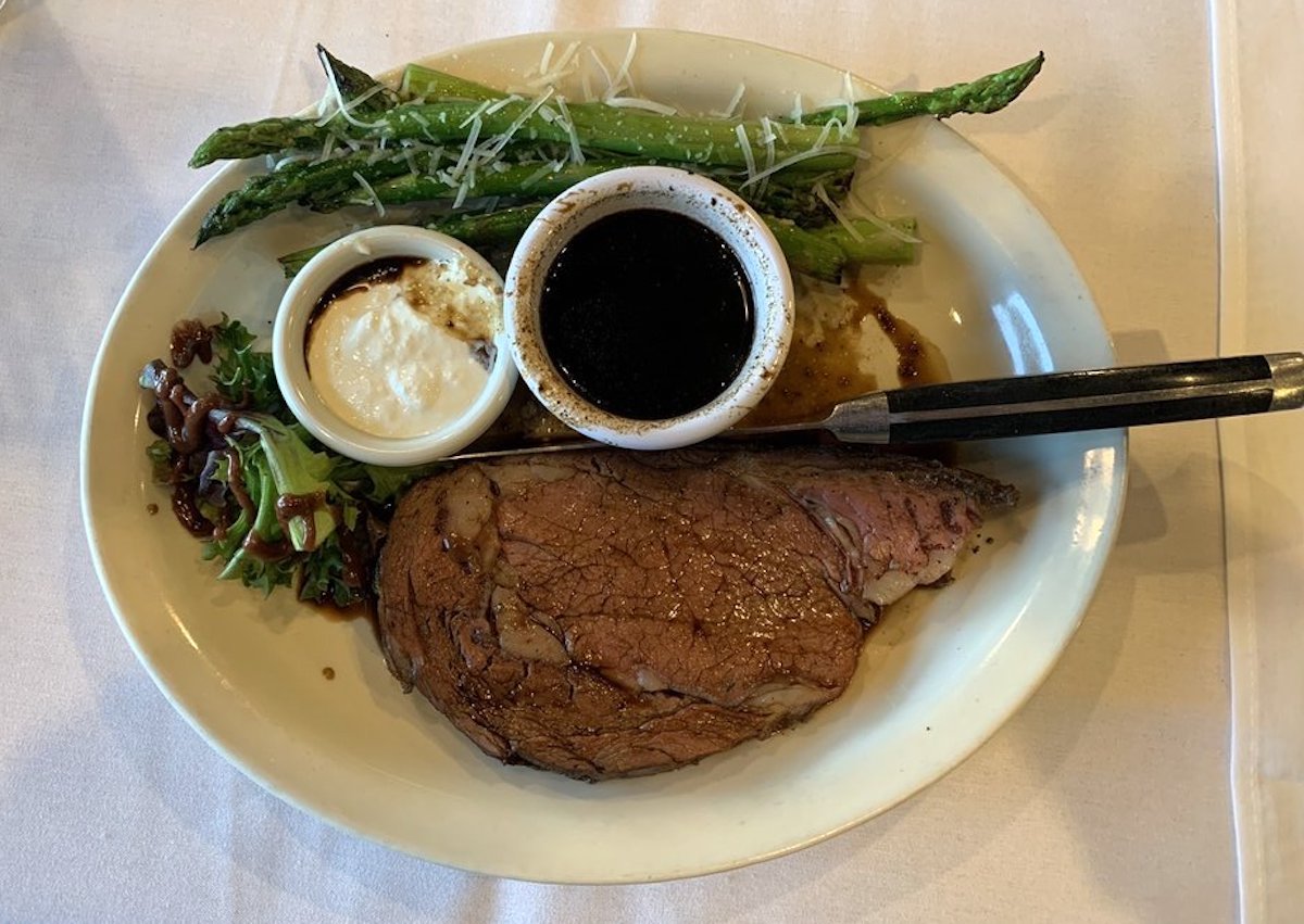 A plate with a slice of prime rib, two spears of asparagus topped with grated cheese, a small salad, horseradish sauce, and a cup of au jus—classic fare often found at cheap steak restaurants. A knife rests on the plate.