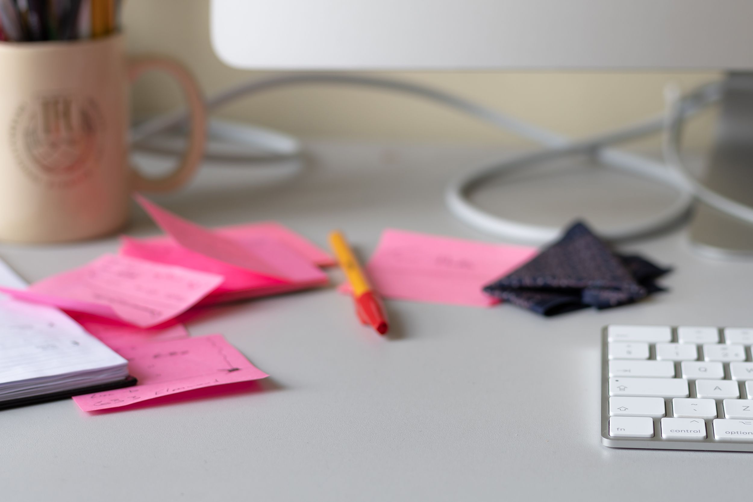computer keyboard, cabling, glasses cloth, diary, red pen, stickie notes, a mug of pens and pencils on a desk