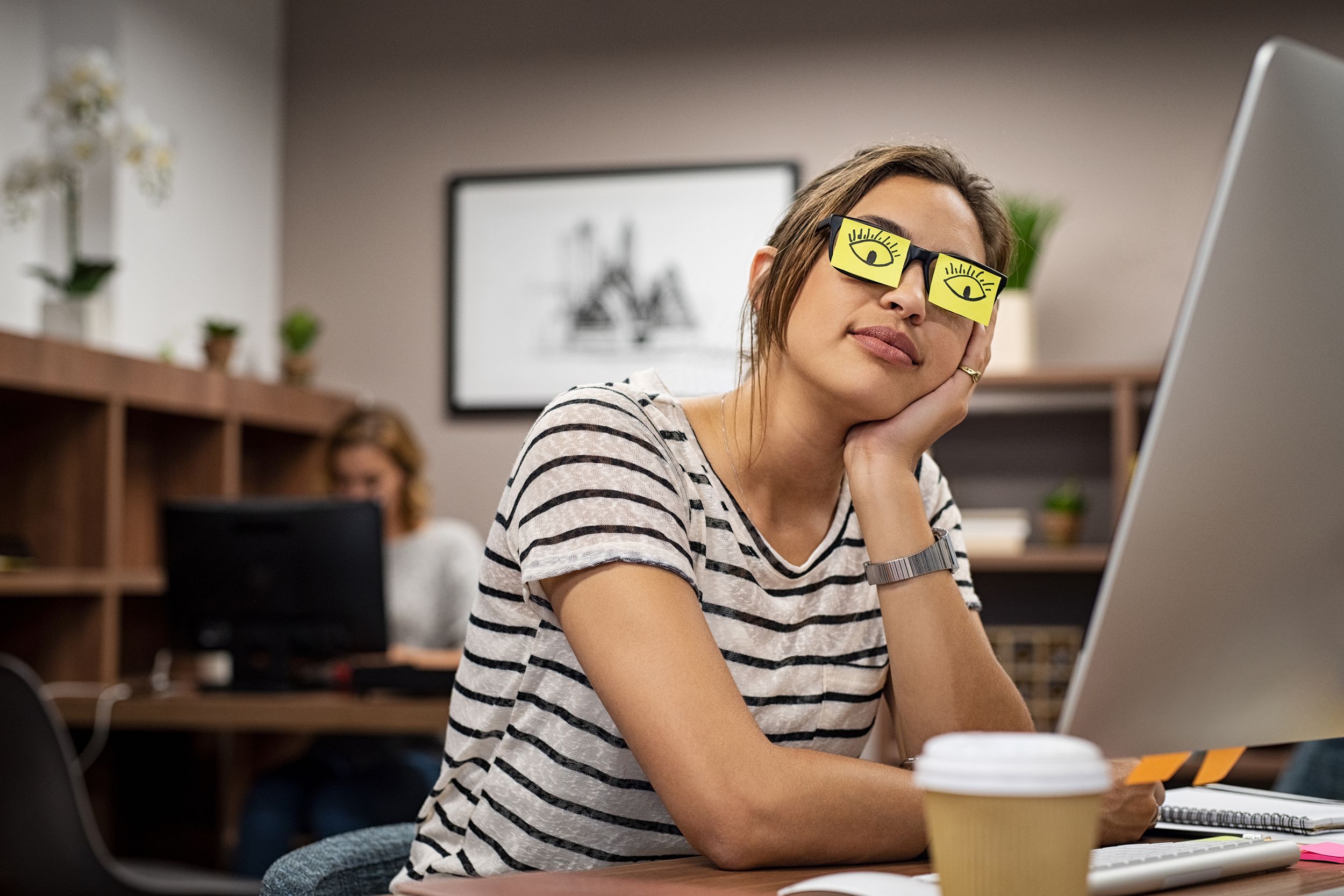 tired woman covering eyes with post it with eyes drawn on them, at the office