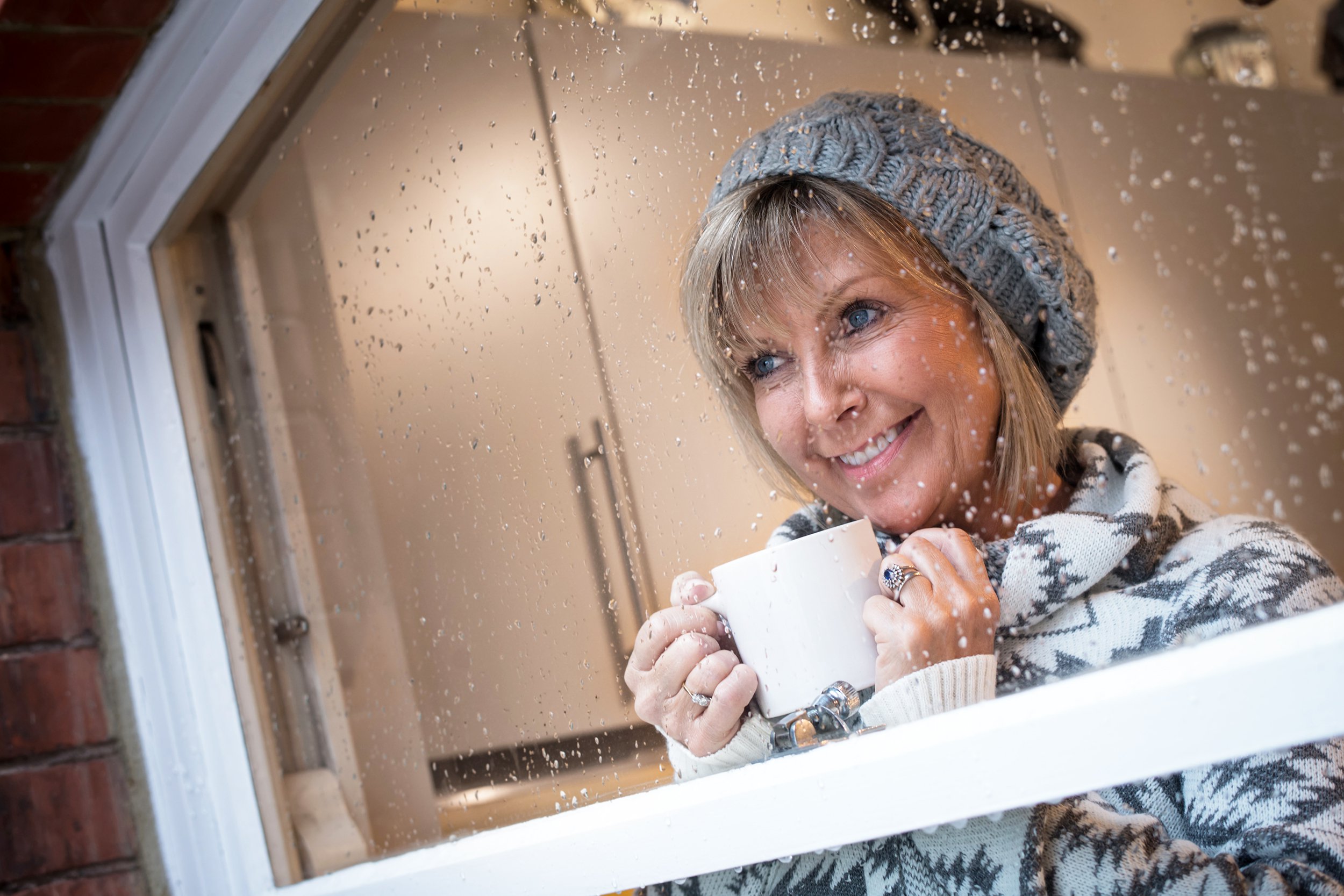 Women looking outside of her rainy window drinking coffee wearing warm clothing