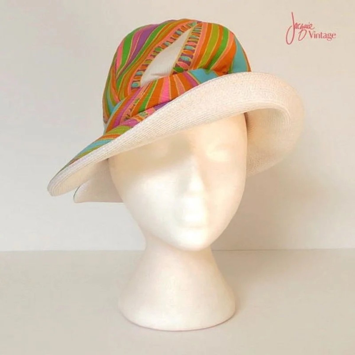 A white mannequin head displays a vintage hat—possibly reminiscent of first Walmart store items—with a wide, white brim and a colorful, diagonally-striped crown in green, orange, pink, and blue. The background is plain and light-colored.