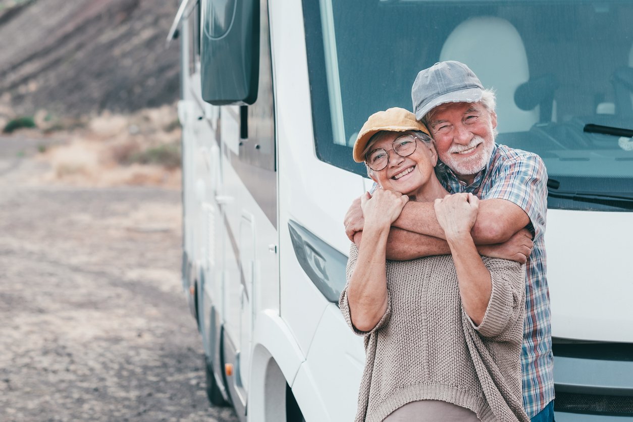 An elderly couple smiles and hugs in front of a white camper van, both wearing casual clothes and hats, enjoying a road trip in a scenic outdoor setting.
