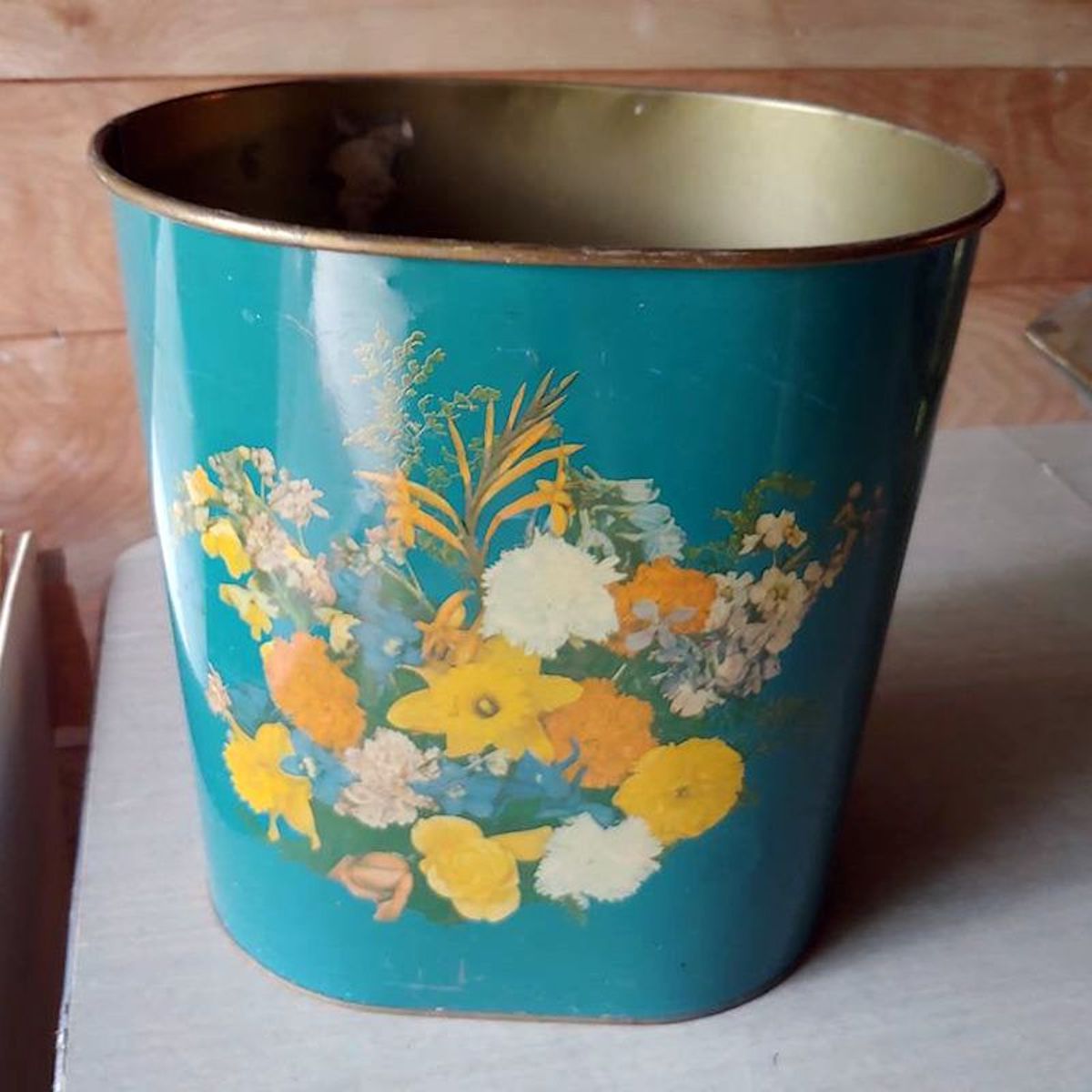 A teal-colored metal wastebasket, reminiscent of first Walmart store items, is decorated with a colorful floral design of yellow, white, and orange flowers, sitting on a light-colored surface against a wooden wall background.