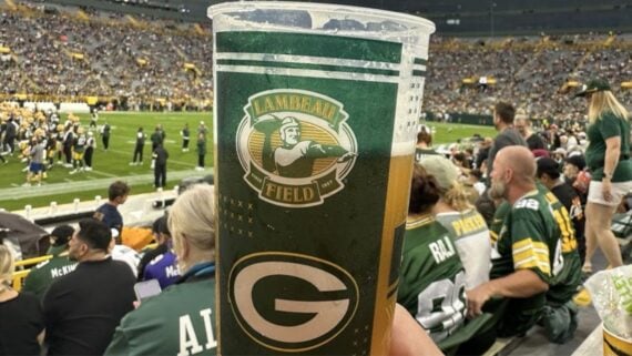 A hand holds a beer cup with "Lambeau Field" and a Green Bay Packers logo in front of a packed stadium, highlighting fans and players, as NFL fans often compare beer and hot dog prices across different stadiums.