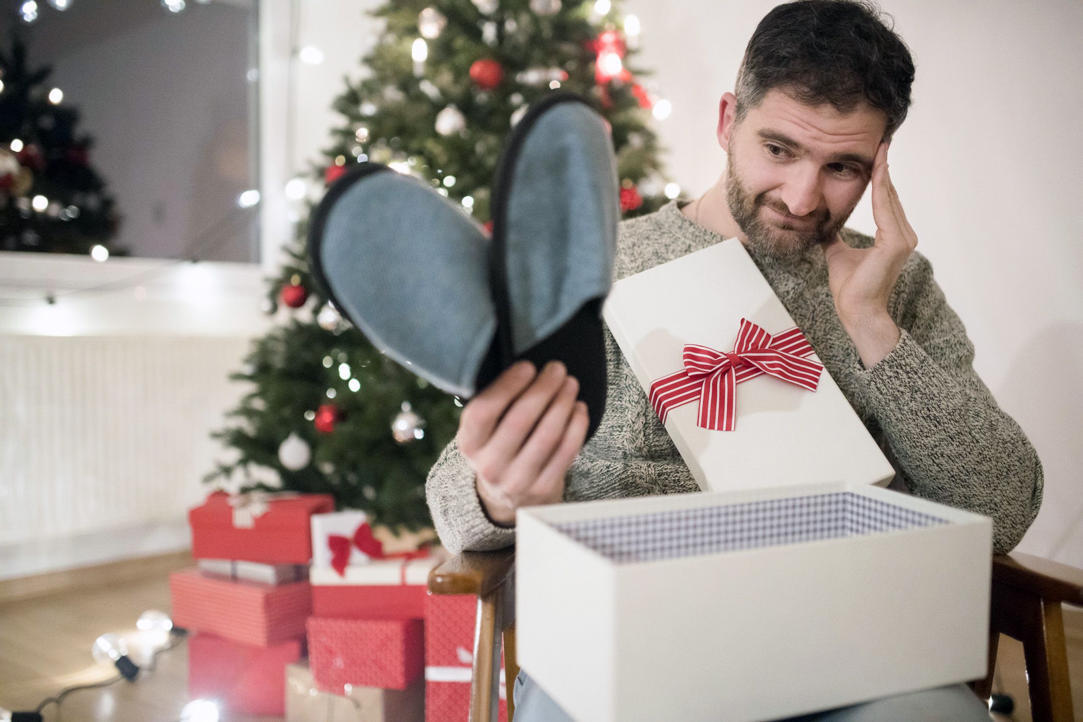 man looking sadly at slippers he has just opened as christmas gift, tree in background
