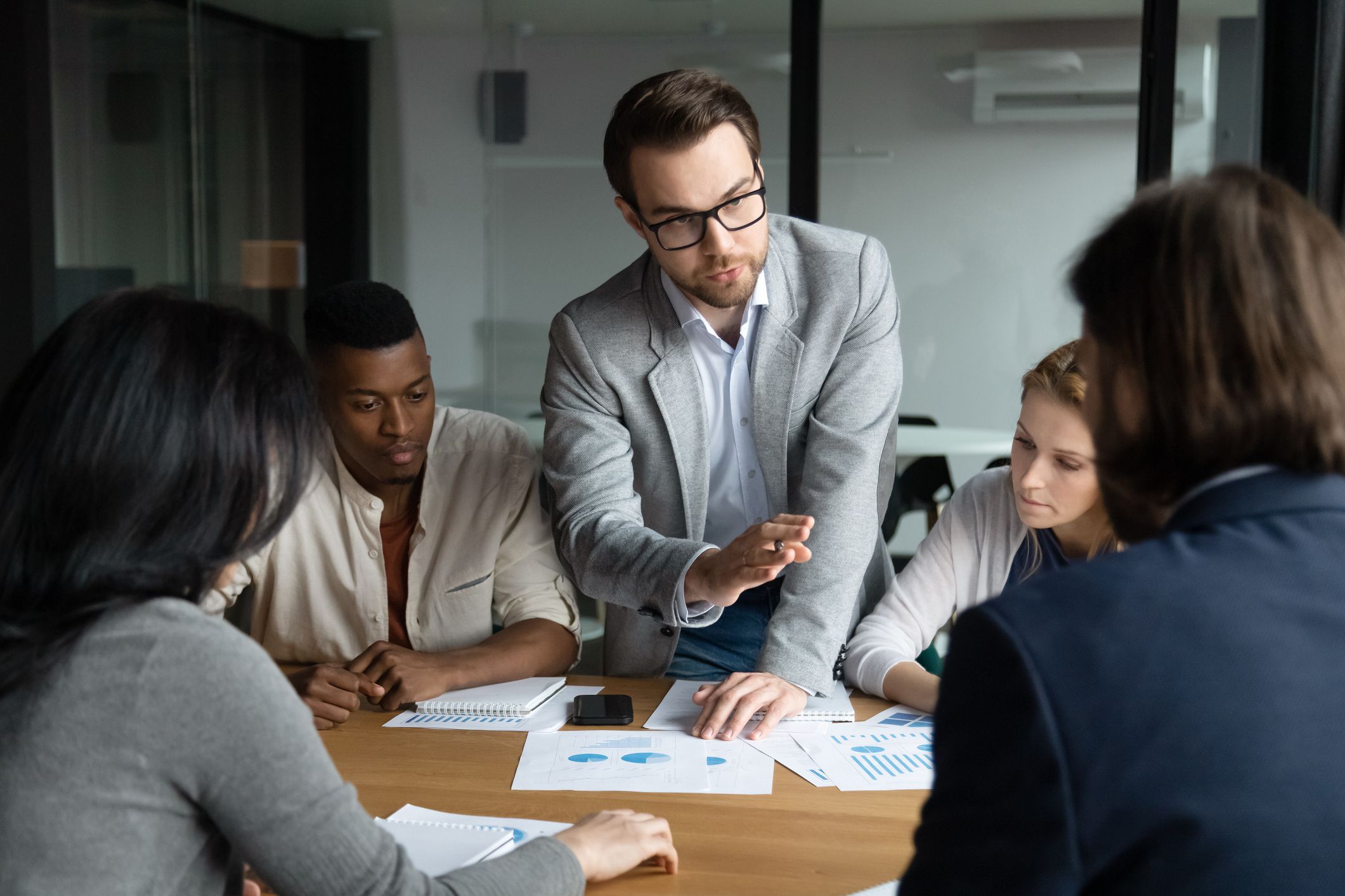 Focused confident young employee in eyeglasses giving arguments about marketing strategy or sharing own business ideas with focused motivated mixed race workgroup at brainstorming meeting in office.