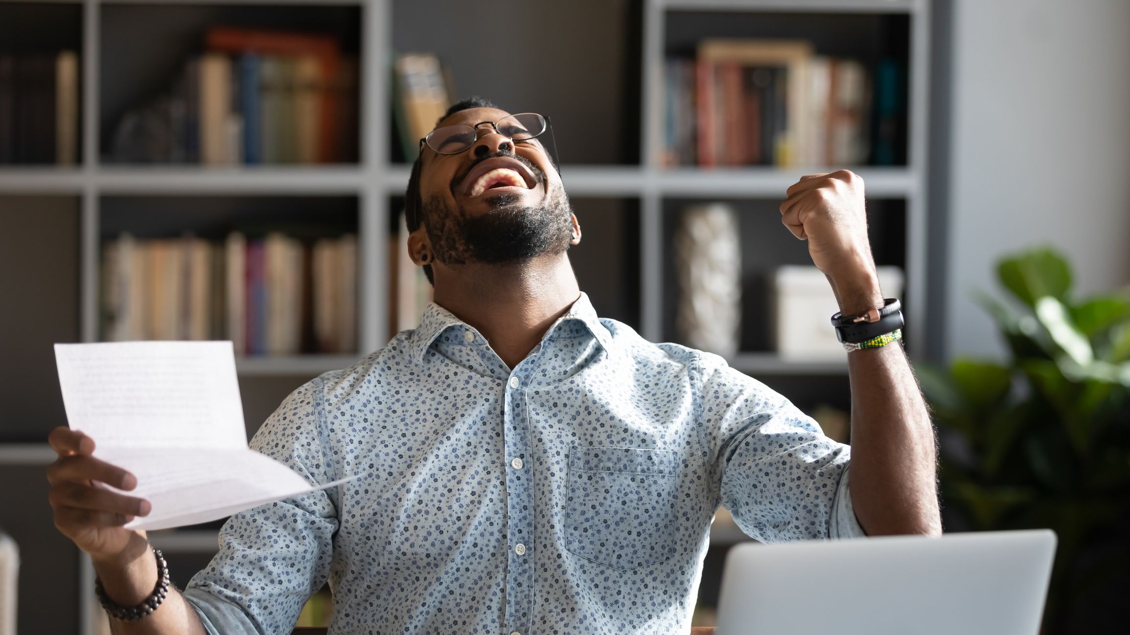 Overjoyed young mixed race man in glasses holding paper in hands, celebrating unbelievable triumph, sitting at workplace. Excited happy millennial african american businessman received loan approval.