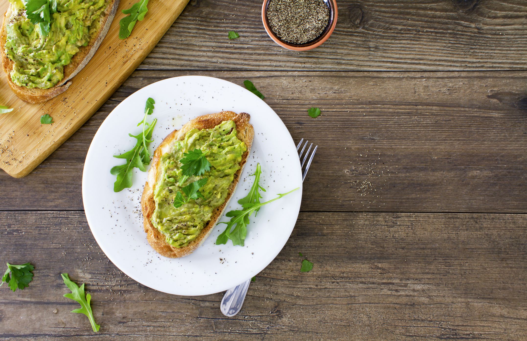 Avocado toast on a white plate on a rustic wooden table. The avocado is sprinkled with chopped parsley and black pepper, and served with fresh arugula. Space for copy on the right of the image. Stock photo.