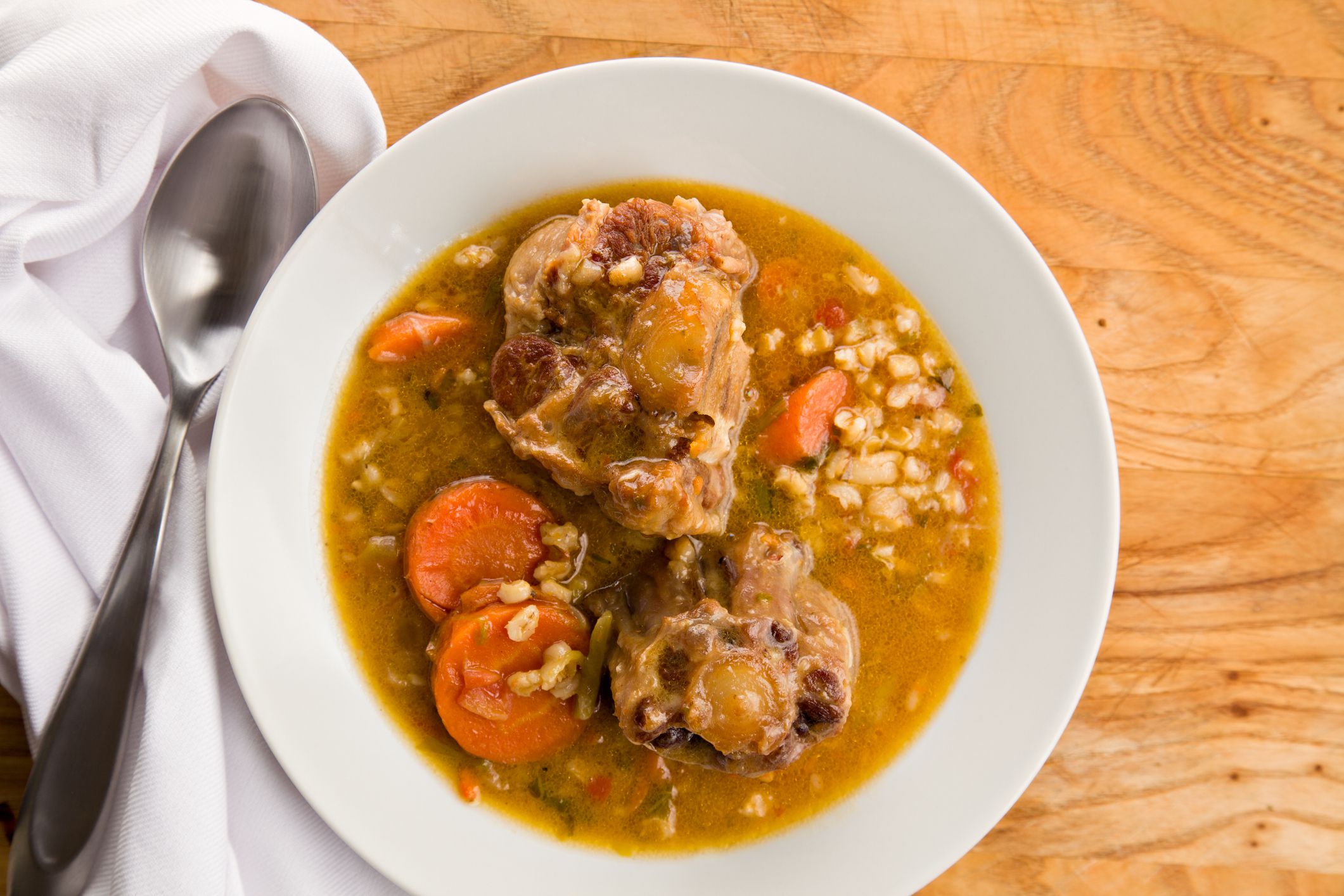 An overhead close up of a bowl of o tail soup a white cloth napkin and metallic spoon sitting on a wooden surface.