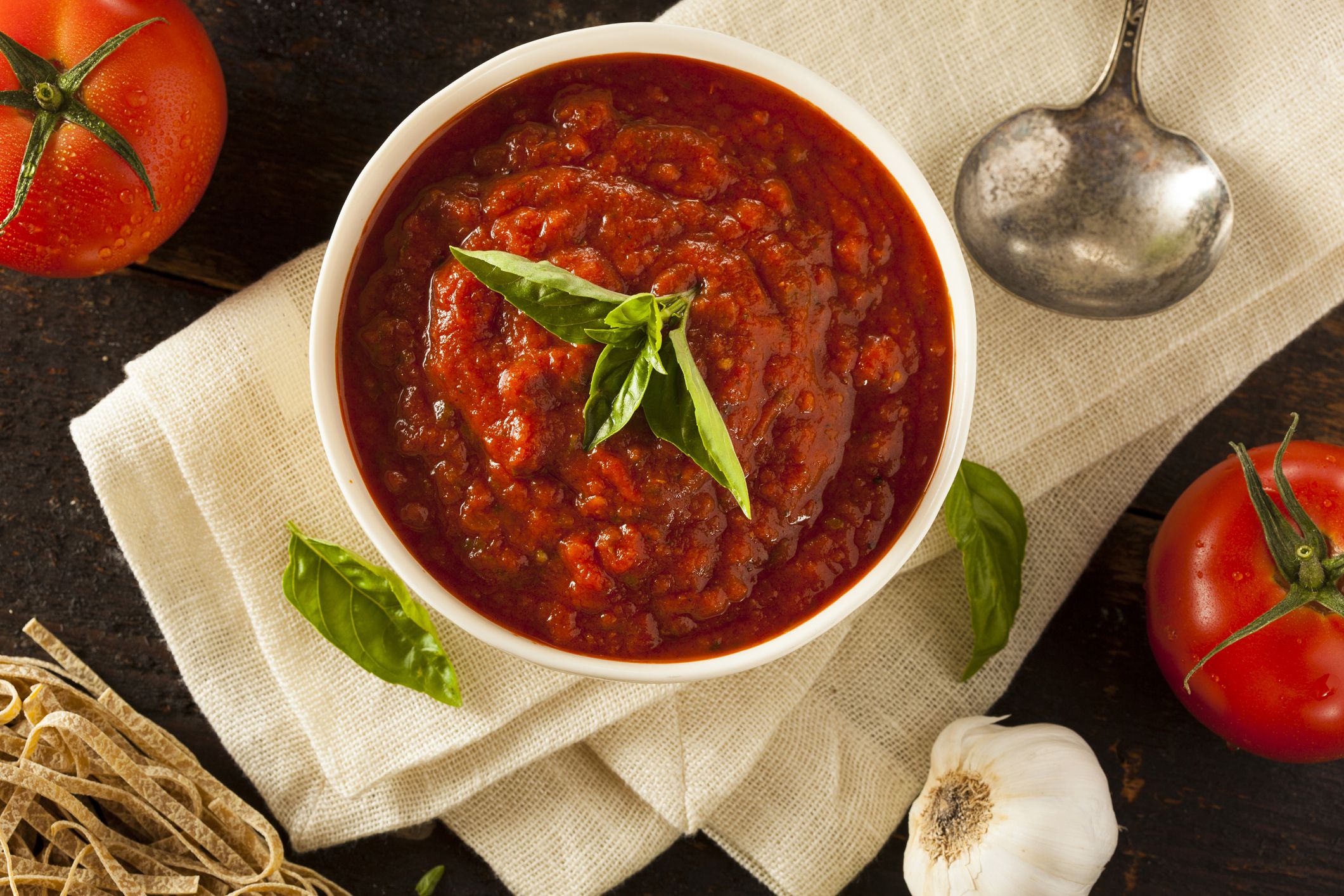 marinara sauce in small dish, spoon, tomatoes, garlic and pasta on side, cloth napkin over black surface