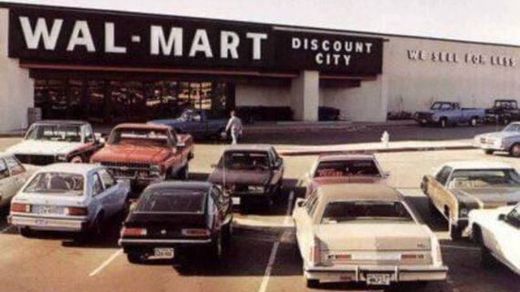 A vintage photo of a Wal-Mart Discount City store with several 1970s and 1980s cars parked in front, evoking the era when first Walmart store items filled its shelves. The building signage reads “WAL-MART DISCOUNT CITY” and “WE SELL FOR LESS.”