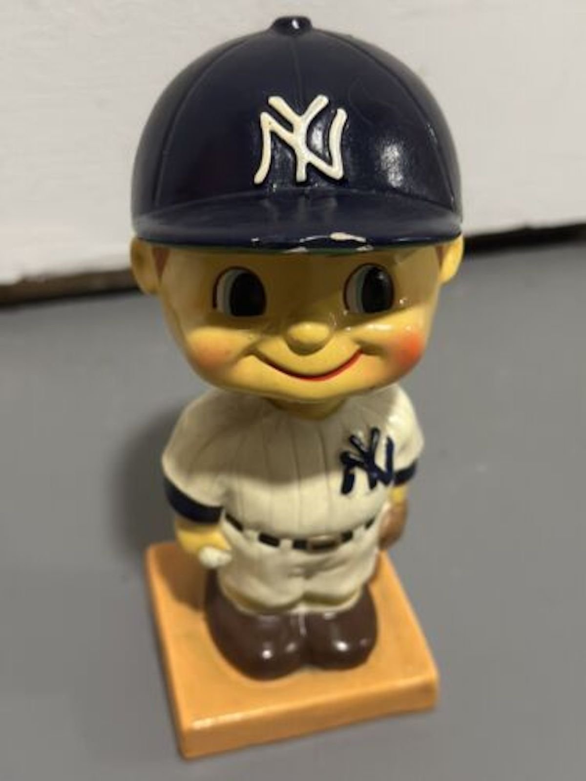A vintage bobblehead figurine of a smiling child in a New York Yankees uniform and cap stands on a square orange base—an appealing piece for fans of expensive collectibles.