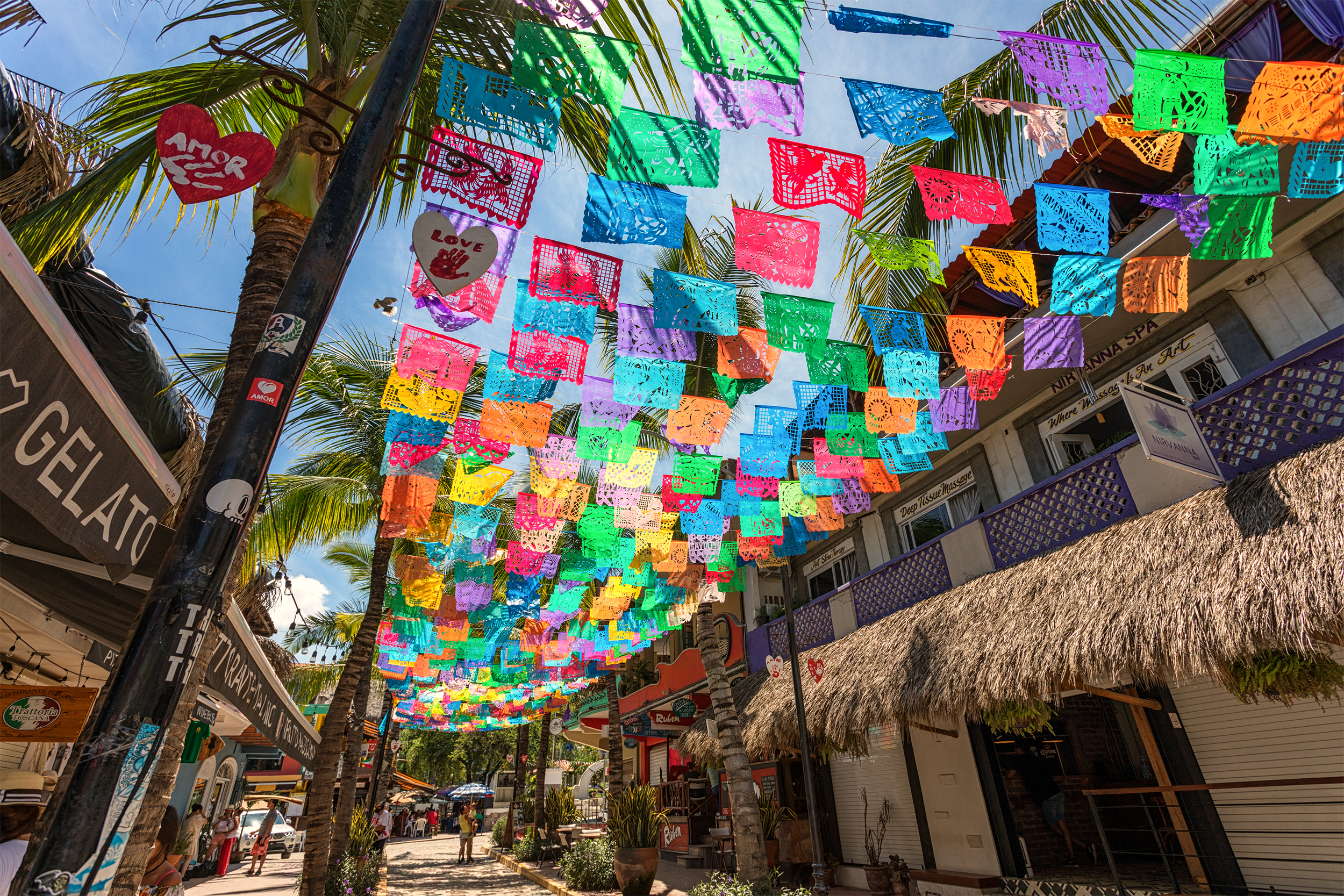 Colorful flags line top of the street in the town of Sayulita, Mexico, looking up, shop buildings on both sides, sunlight coming through flags and palm tree leaves