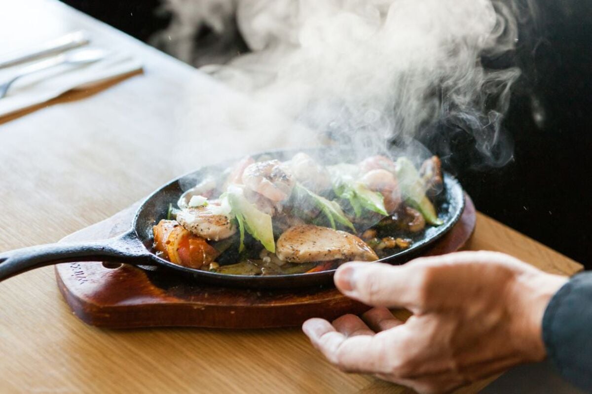 Fish and Shrimp Mexican Dish on a cast iron skillet on a wooden holder, La Serenata de Garibaldi, Los Angeles, selective focus, male’s left hand just sit it down on a wooden table at the restaurant blurred