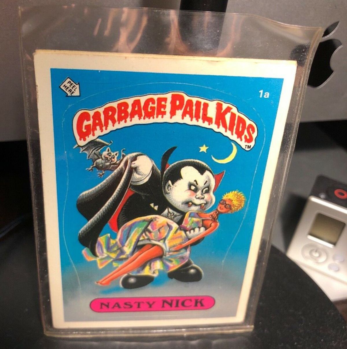 A Garbage Pail Kids trading card titled "Nasty Nick" shows a cartoon vampire holding a doll, with a small bat flying nearby. Known among expensive collectibles, the card is protected in a sleeve on a dark surface.