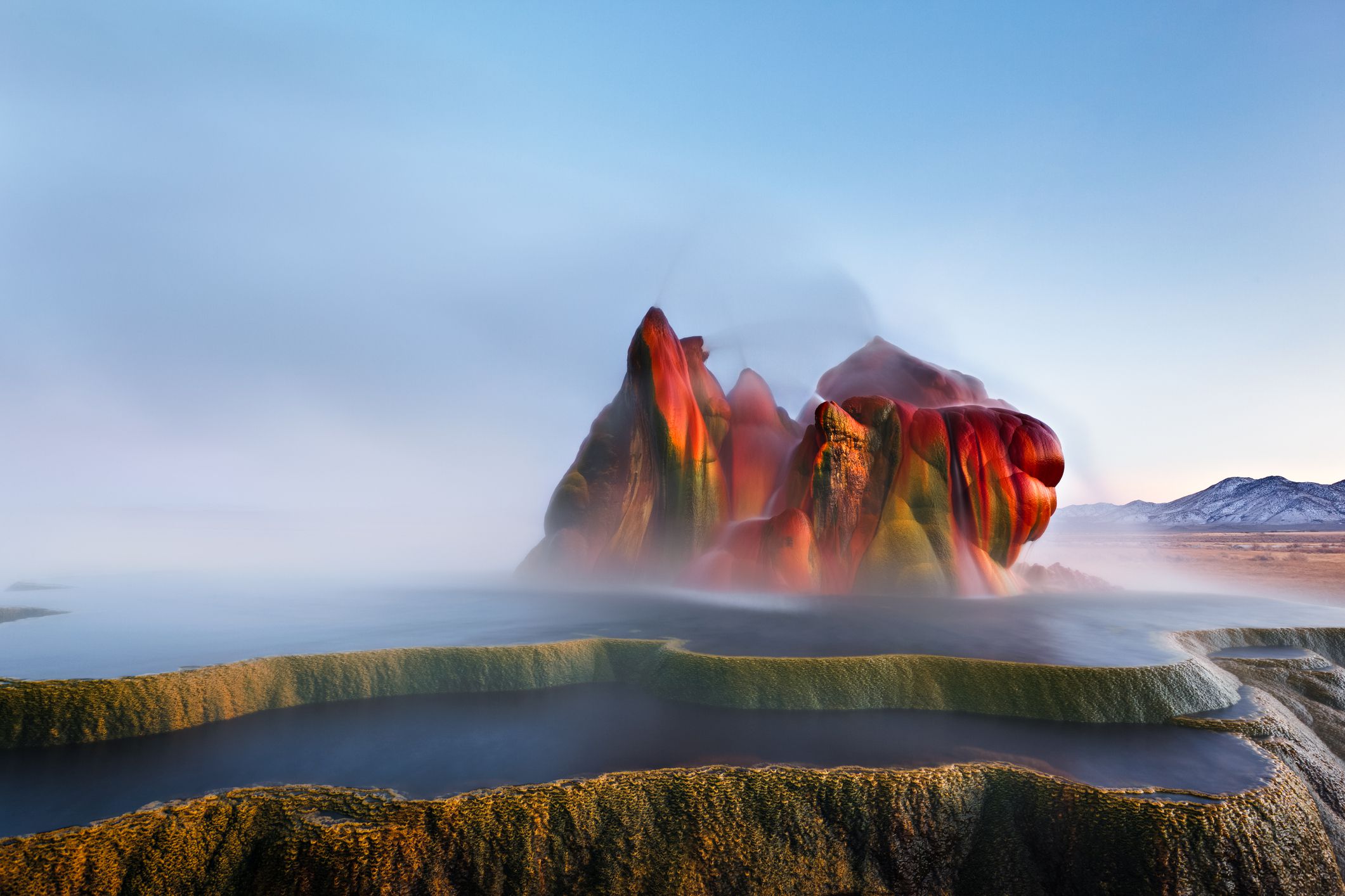 Colorful mineral formations of Fly Geyser rise from steaming, terraced pools in a Nevada desert landscape under a clear sky, with mountains visible in the background.