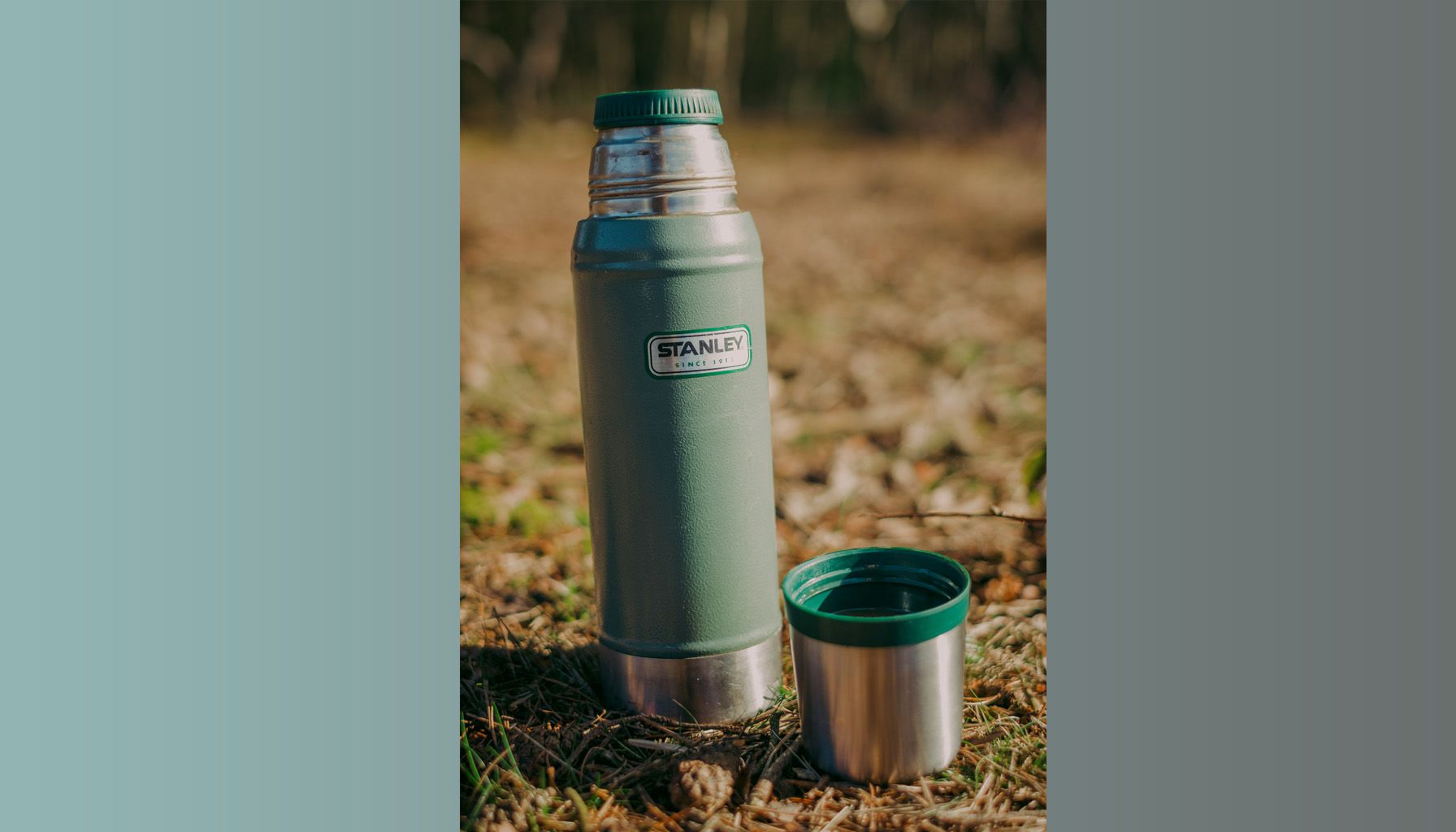 A vintage Stanley thermos in hammertone green is shown in the grass in an outdoorsy setting.
