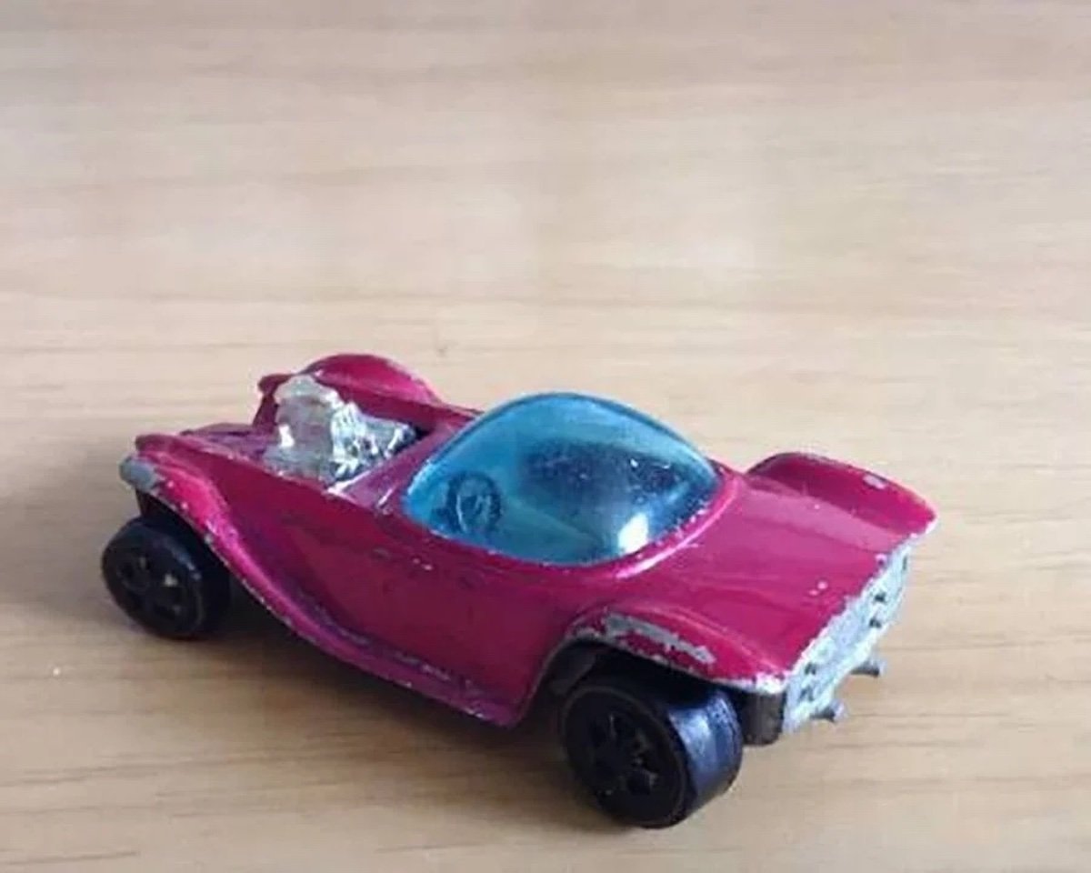 A small, worn red toy car with black wheels, a blue-tinted windshield, and a visible silver driver figure sits on a light wooden surface—once ordinary, now coveted among expensive collectibles.