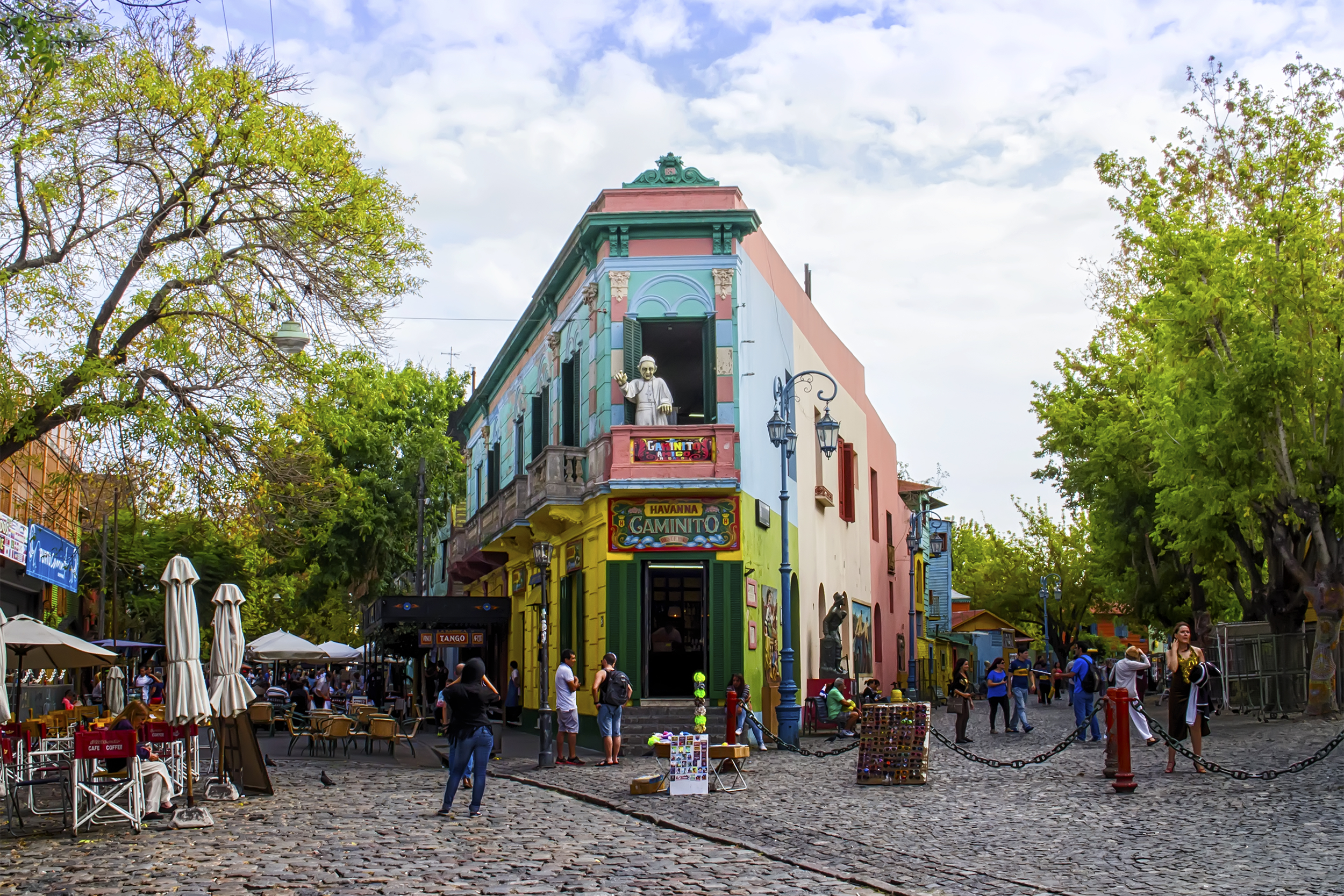 La Boca, Buenos Aires, Argentina, colorful building in the center, surrounded by green trees, people walking on cobble stone streets, on a sunny day, white clouds in the blue sky