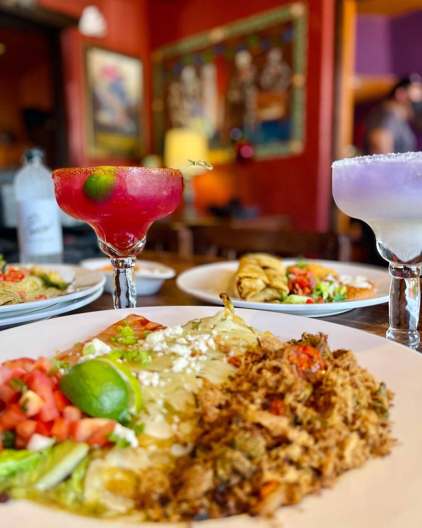 Closeup of many dishes and two Margaritas on a wooden table at the restaurant, El Charro Café, Tucson, colorful walls and paintings are blurred in the background