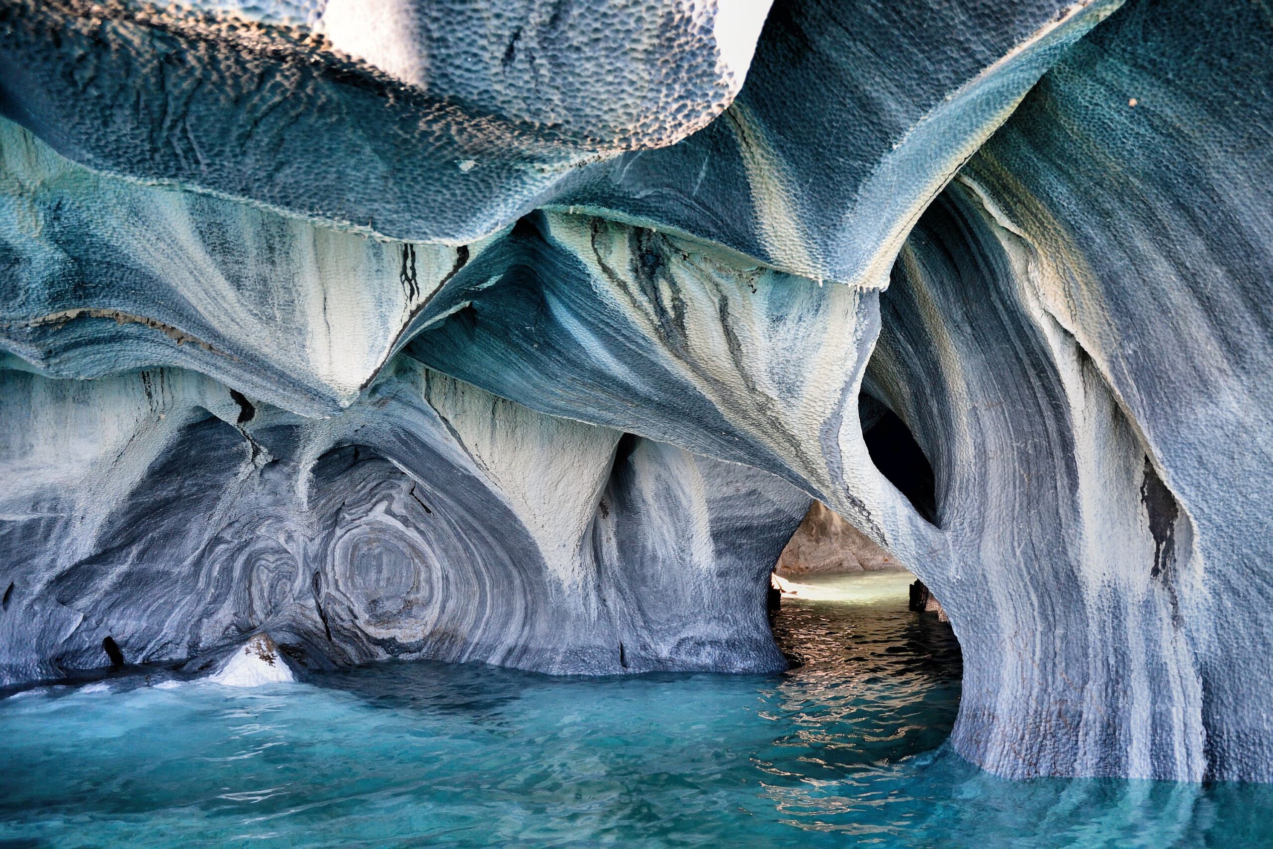 Blue-green water flows through intricately carved marble caves with smooth, swirling gray and white stone formations, creating dramatic natural arches and openings shaped by erosion.