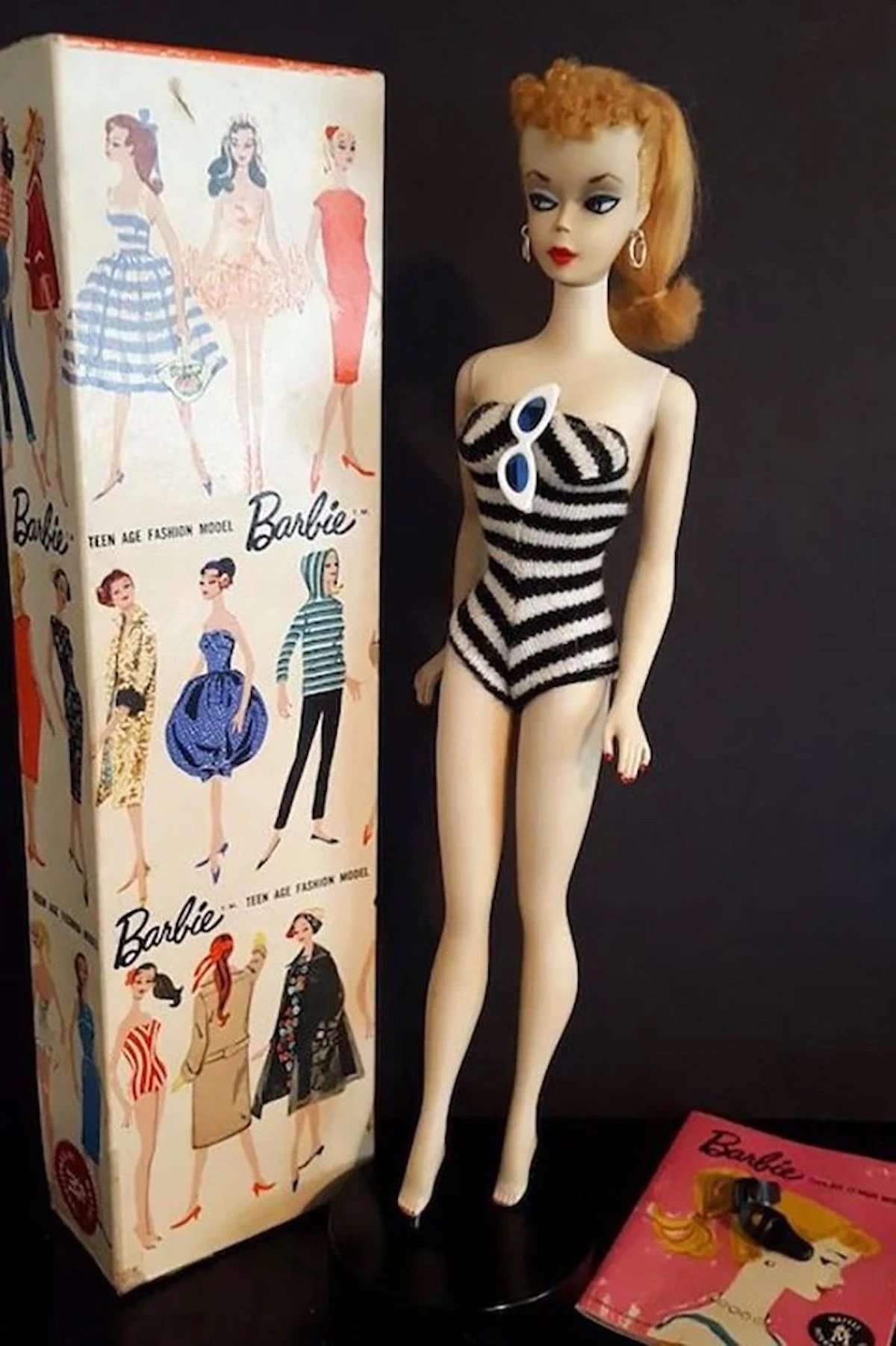 A vintage Barbie doll, prized among expensive collectibles, stands in a black-and-white striped swimsuit next to its original box adorned with illustrated Barbie designs, a pink booklet, and classic accessories like blond hair, hoop earrings, and sunglasses.