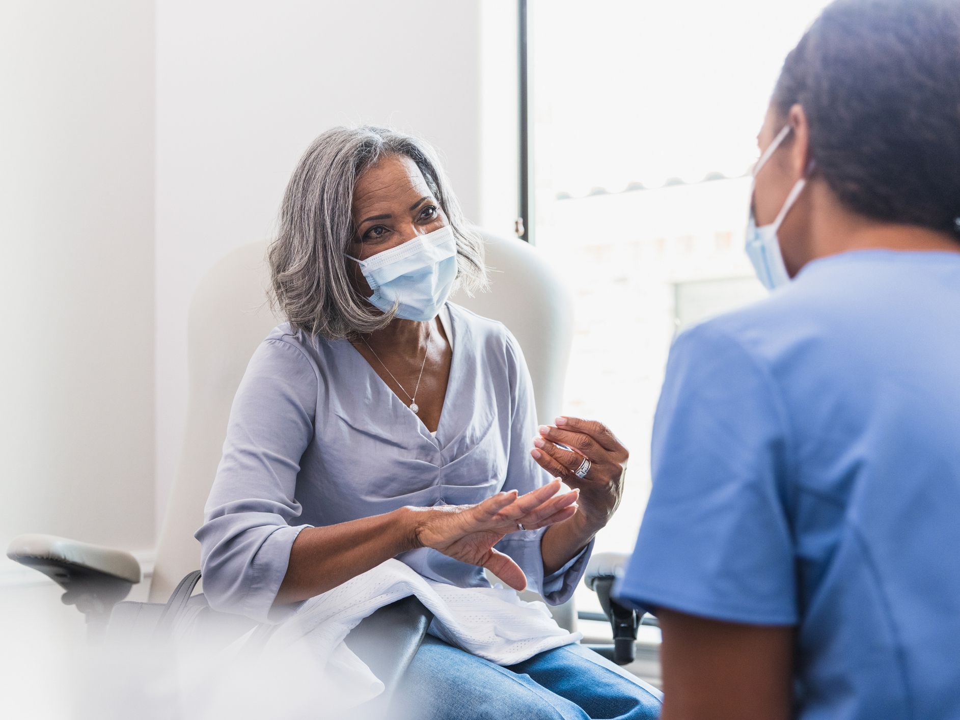 A senior woman, wearing a protective face mask, talks with a female nurse during a medical appointment.
