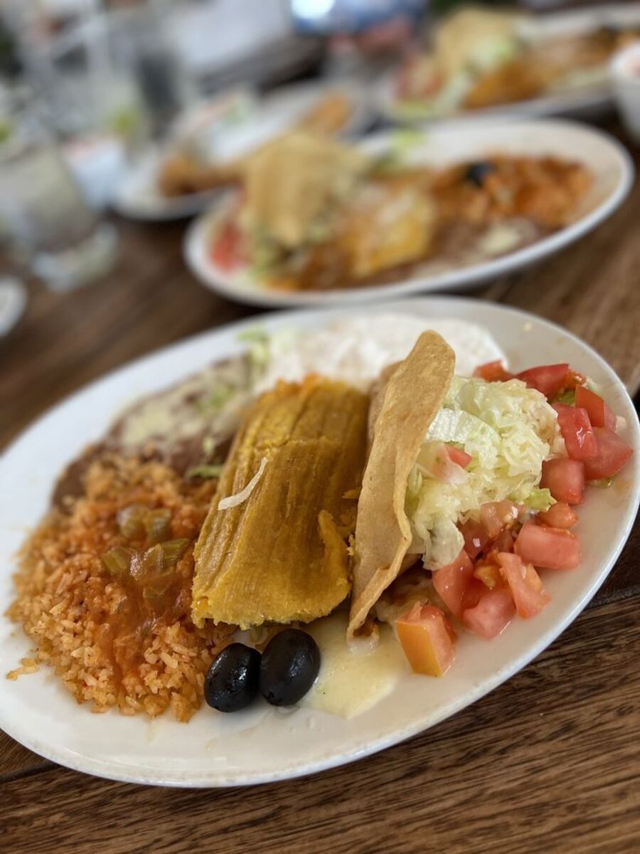 Tacos and Sweet Corn Tamale with sides on a white ceramic plate, Casa Vega, Sherman Oaks, California, selective focus, three additional meals on plates with glasses of drinks on a wooden table at the restaurant