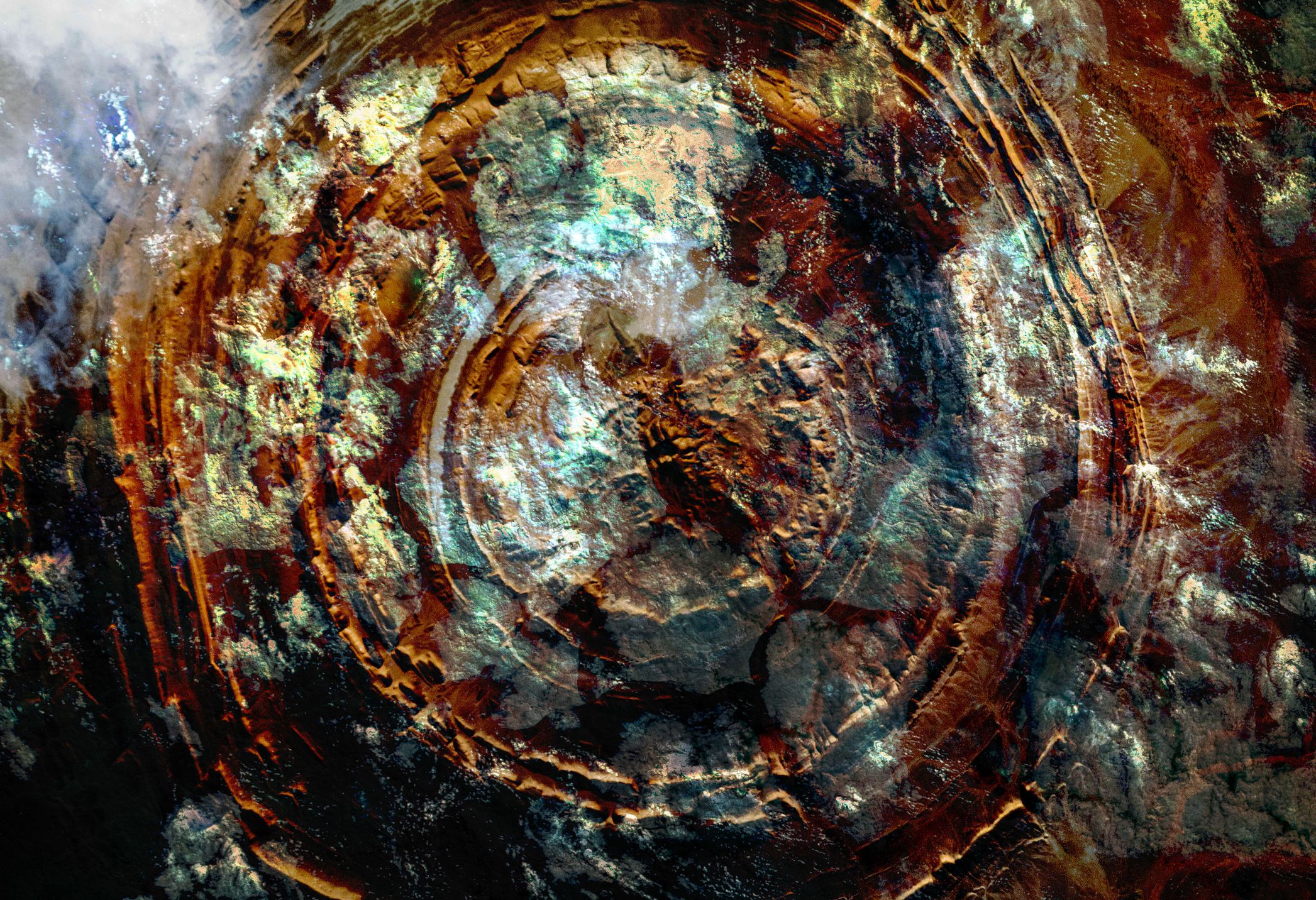 Aerial view of the Richat Structure in Mauritania, showing concentric circular rock formations in shades of brown, gold, and blue-green, with clouds partially covering the upper left corner.