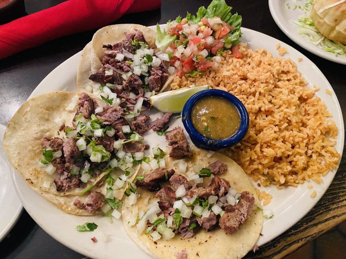 Three Steak Street Tacos with sides on a white plate, El Fenix, Dallas, with utensils rolled in a red napkin and another dish on a wooden table at the restaurant