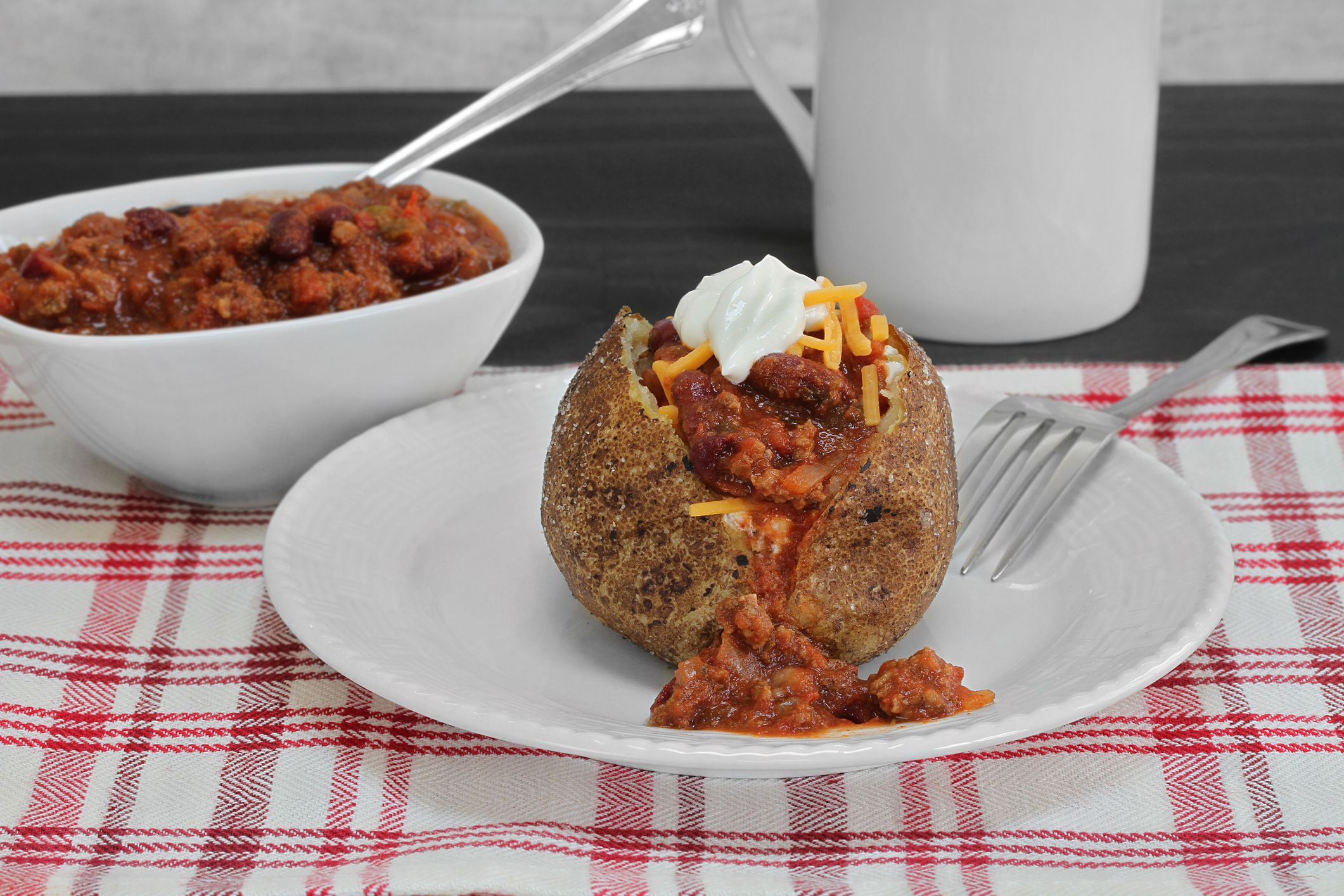 One baked potato topped with chili, cheddar cheese and sour cream. Macro, close up. Background includes a bowl of chili.