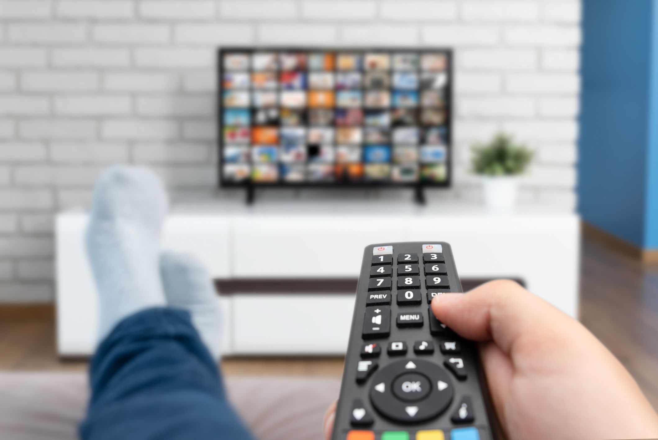 Man watching TV, lying on sofa, legs on table. Person holding remote control in living room