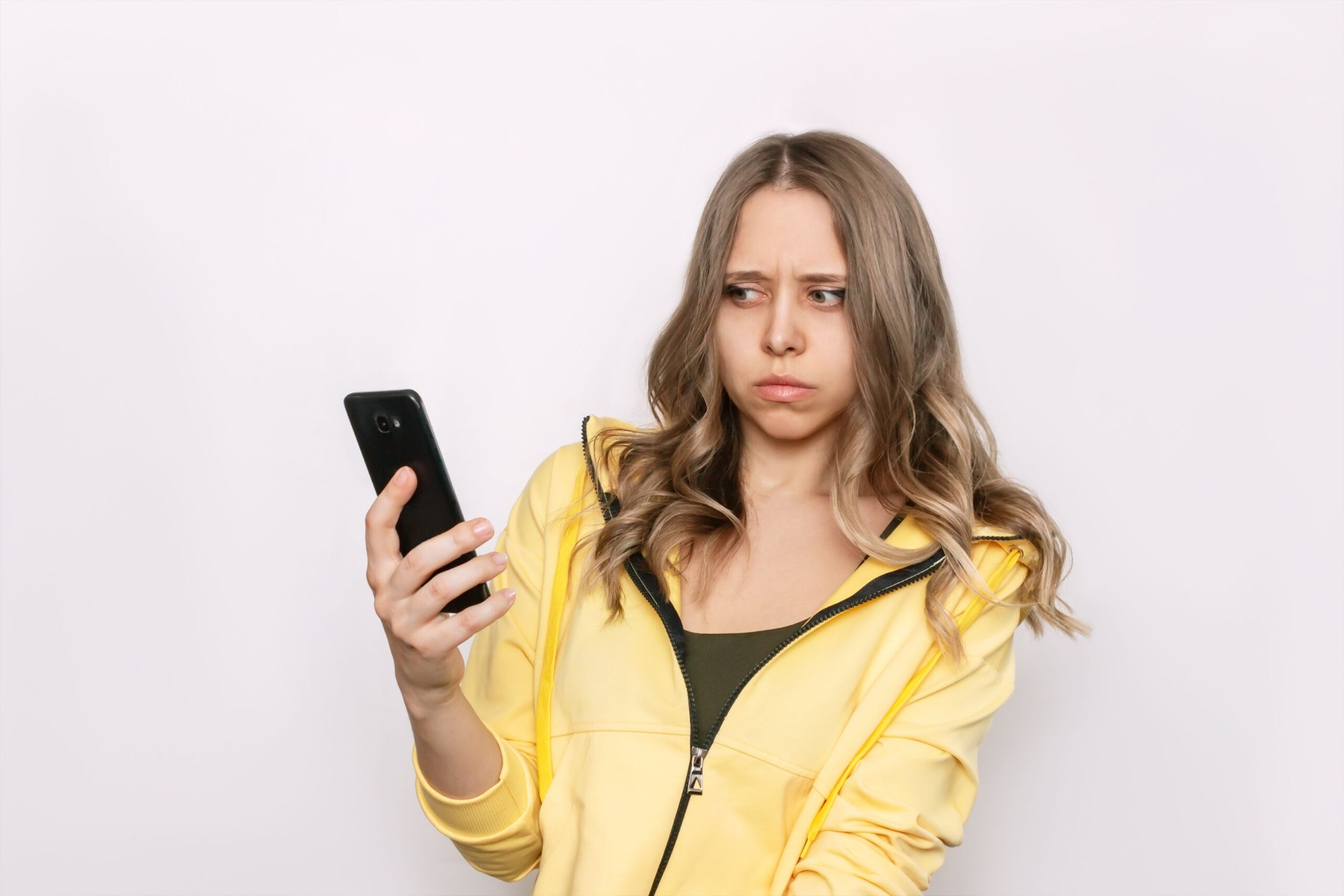 A frightened young caucasian blonde woman with wavy hair holds a mobile phone in her hand looking suspiciously at the incoming call from an unknown number isolated on a white background. Fraud