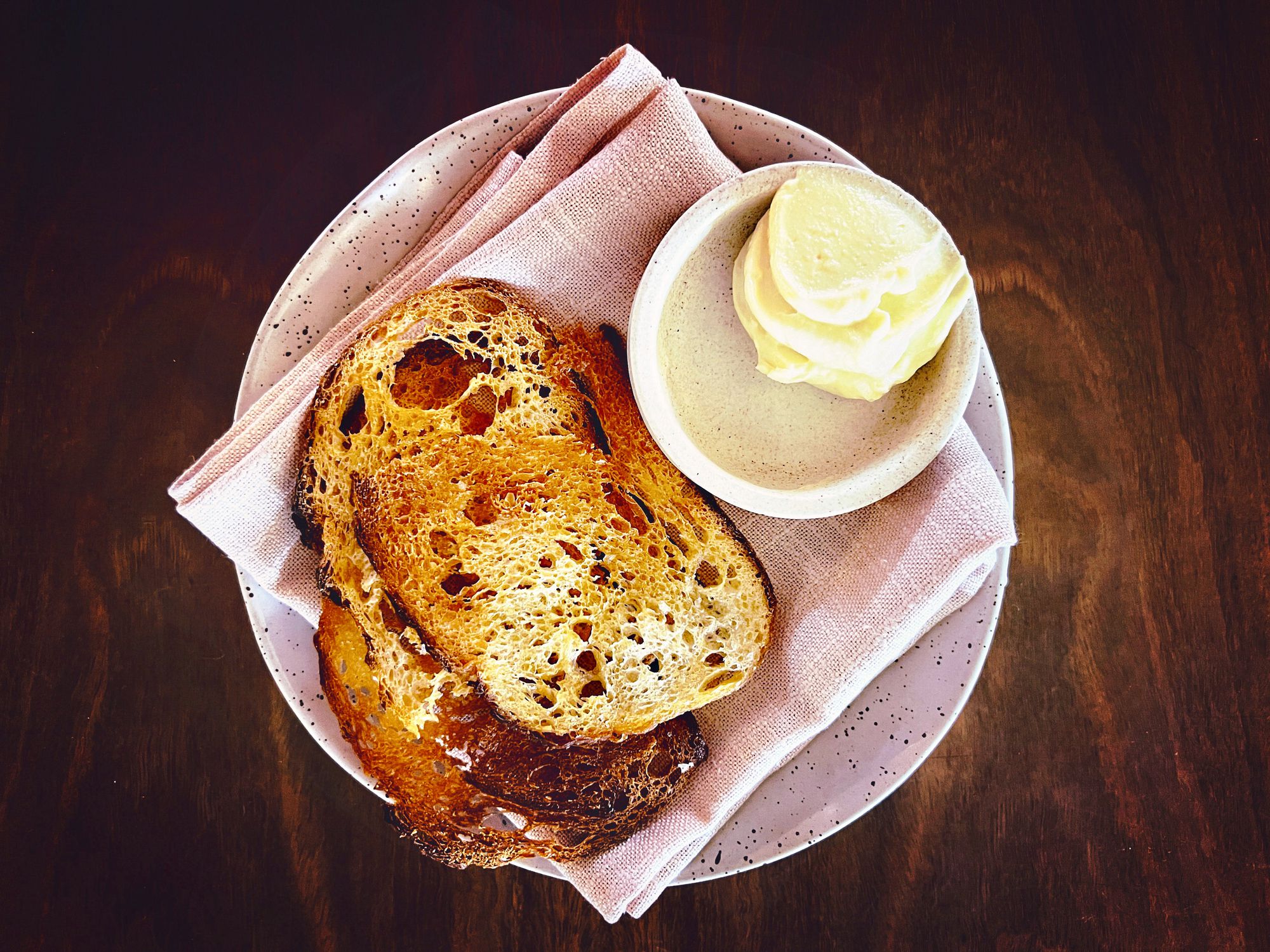 Horizontal high angle closeup photo of two slices of toasted house made sourdough bread with cultured butter in a small white bowl on a linen napkin on a handmade ceramic plate in a fine dining restaurant. Byron Bay, NSW.