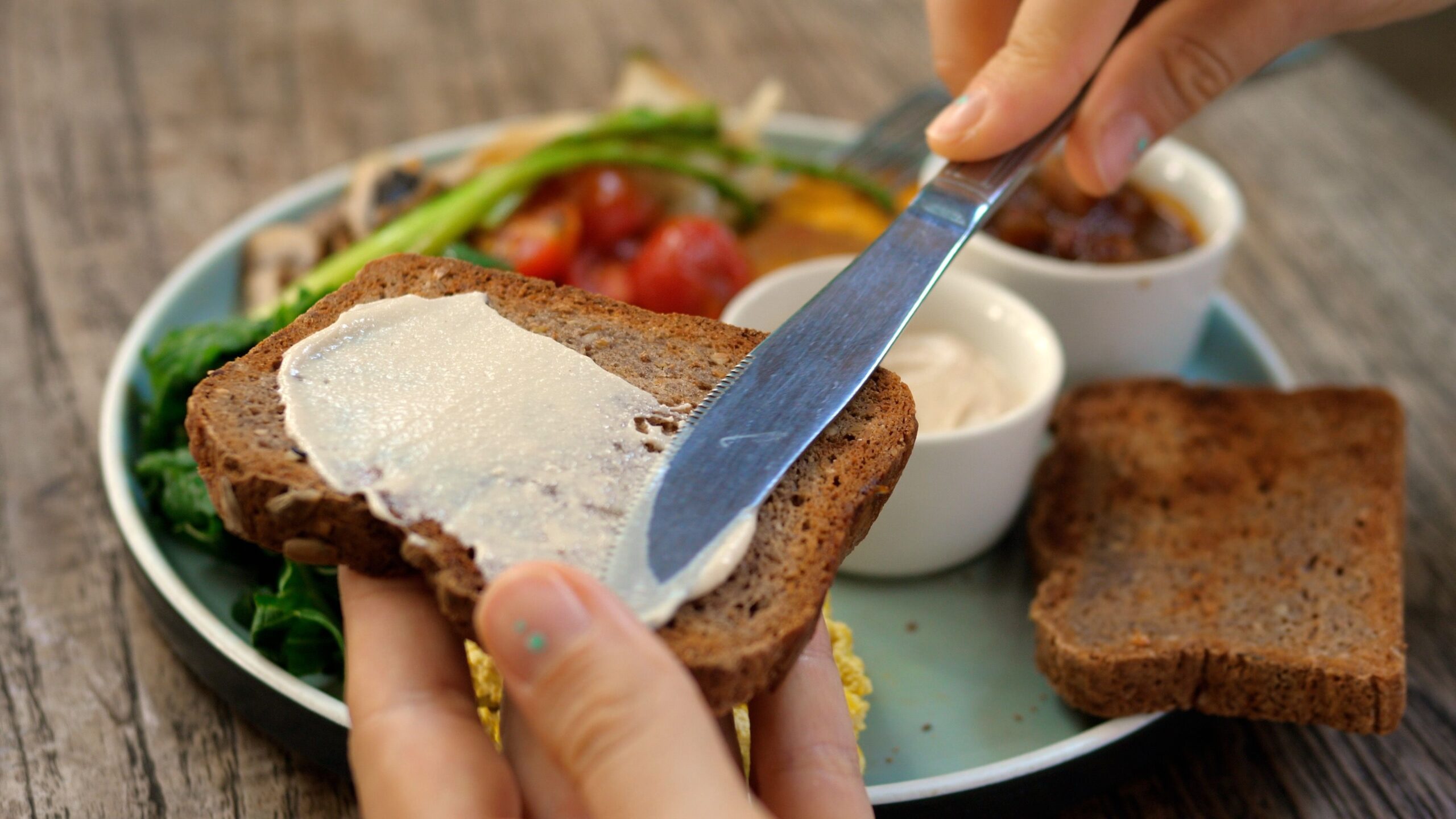 Breakfast in the fresh air in your favorite cafe. A plate with vegetables and tofu for a complete diet. Close-up of a knife spreading sauce on a piece of whole grain toast.