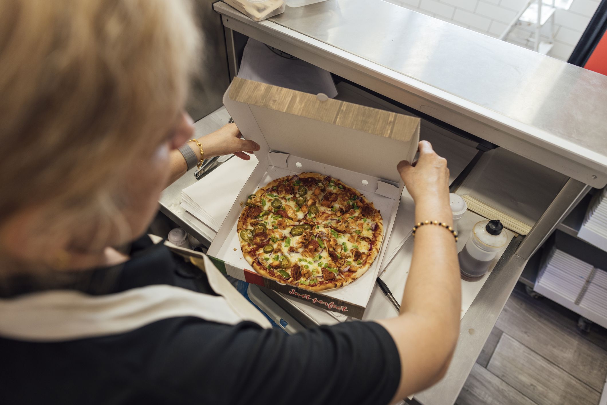 Over the shoulder a mature Indian woman wearing all black casual clothing and an apron closing a pizza box with a freshly made chicken pizza inside, she is working in a family-run fish and chip shop in Gateshead, England.