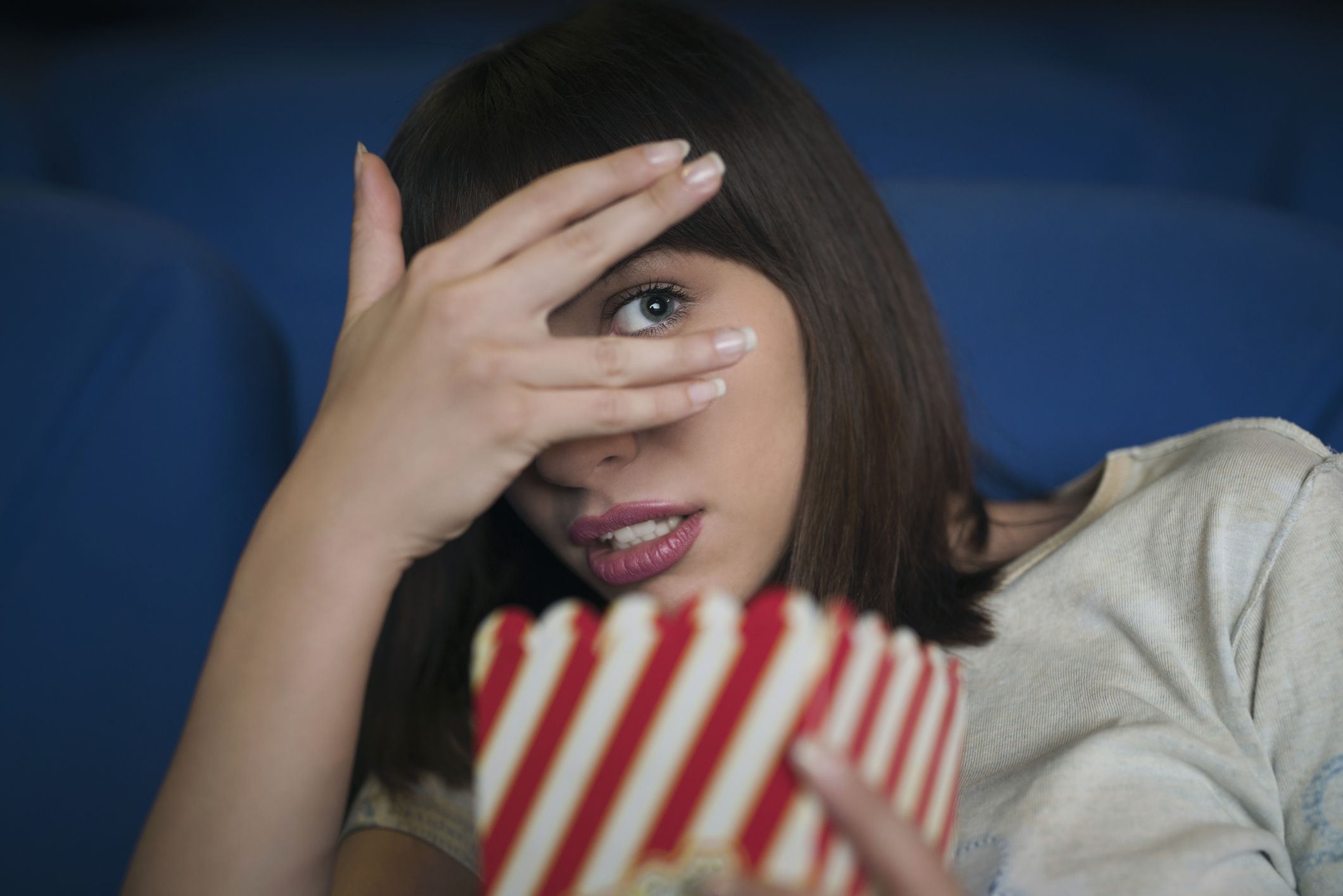 Young woman in the cinema, watching a scary movie, hiding her look with a hand and holding a box with popcorn