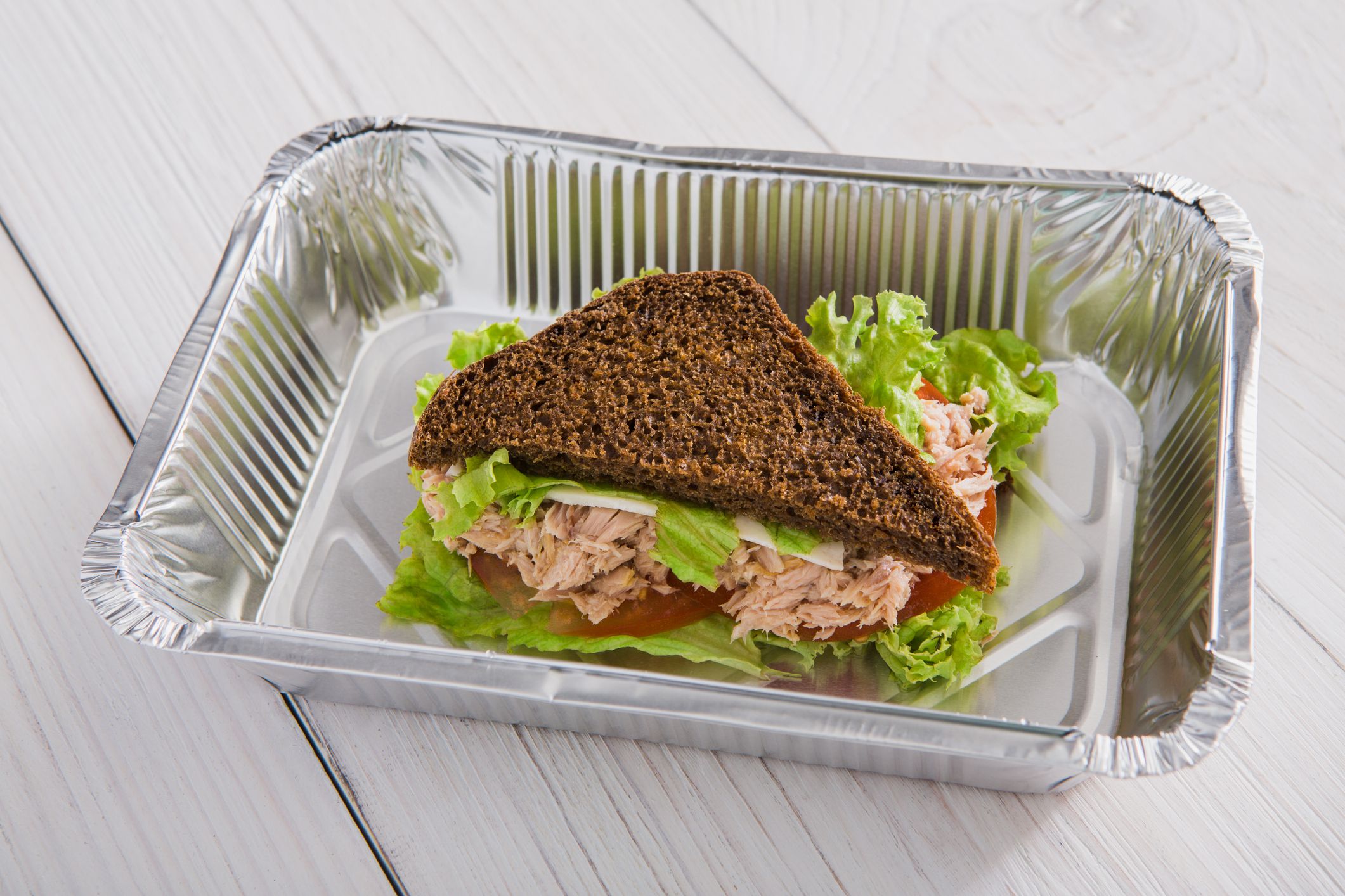 Healthy food delivery and diet concept. Take away of fitness meal. Weight loss lunch in foil box. Whole-grain sandwich with tuna fish and fresh vegetables closeup at white wood