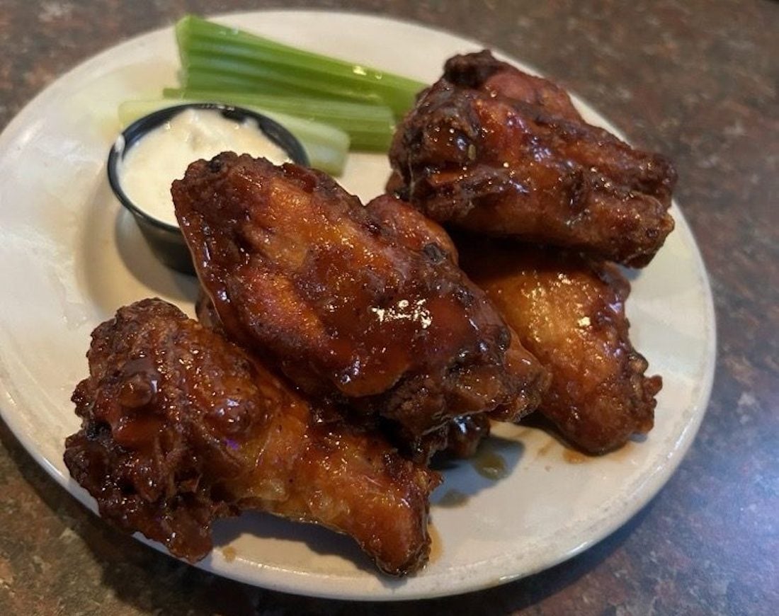 A plate of the best chicken wings glazed to perfection, served with celery sticks and a small cup of ranch or blue cheese dressing on a brown tabletop.
