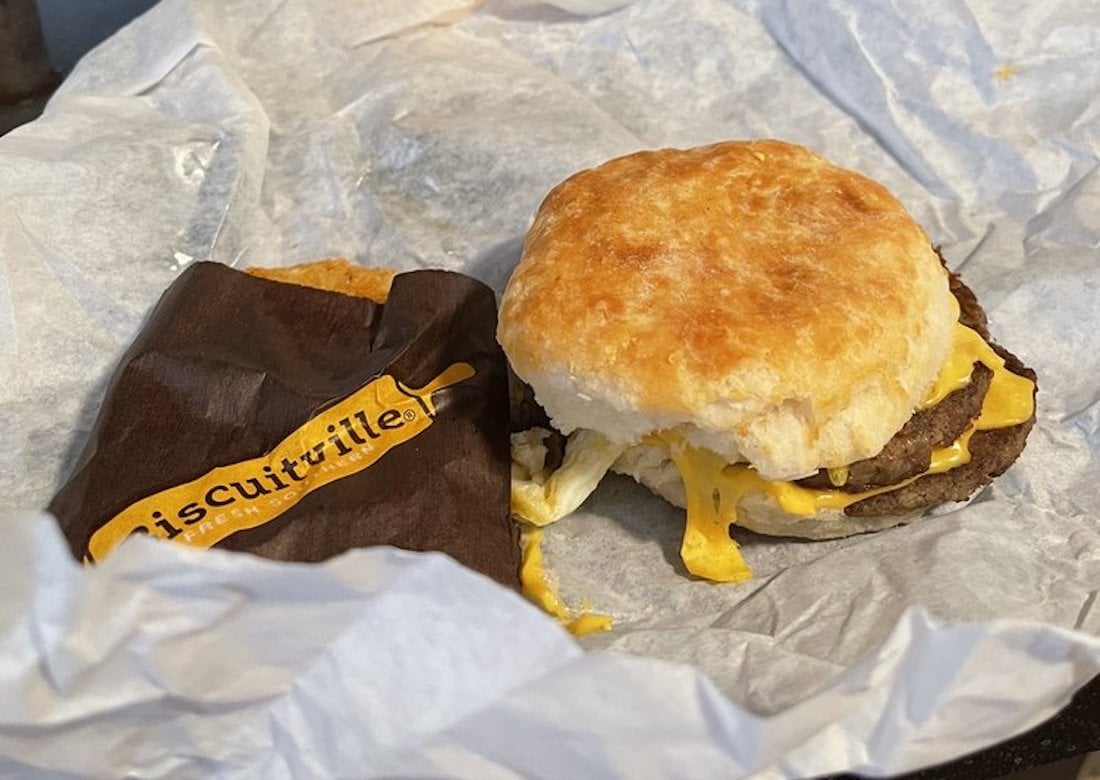 A breakfast biscuit sandwich with egg, cheese, and sausage sits on crumpled white paper next to a partially visible brown wrapper labeled "Biscuitville.