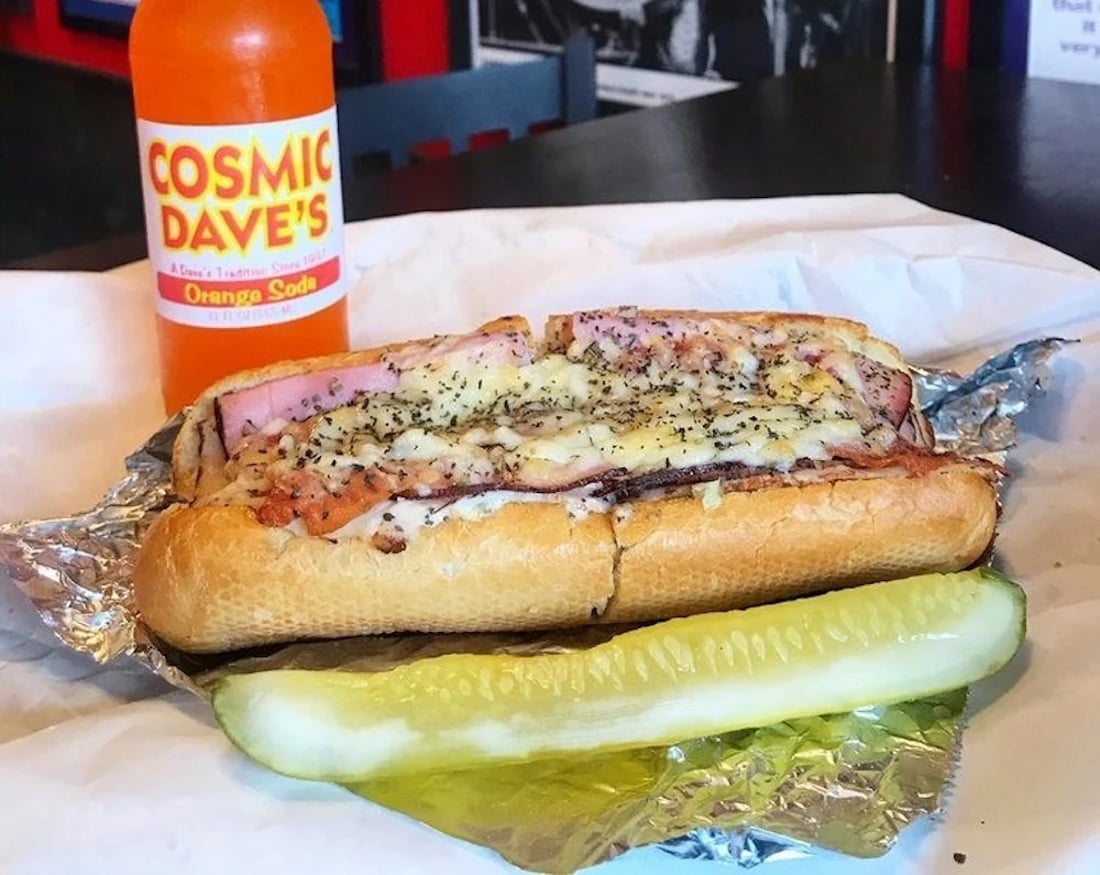 A toasted sub sandwich with melted cheese and ham sits on foil, served with a pickle spear. Behind it is an orange soda bottle labeled "Cosmic Dave’s Orange Soda." The setting appears to be a casual restaurant.