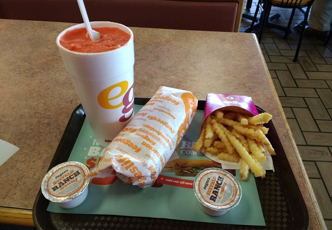 A fast food meal on a tray includes a large red slush drink, a wrapped sandwich, crinkle-cut fries in a pink container, and two cups of buttermilk ranch sauce on a table.